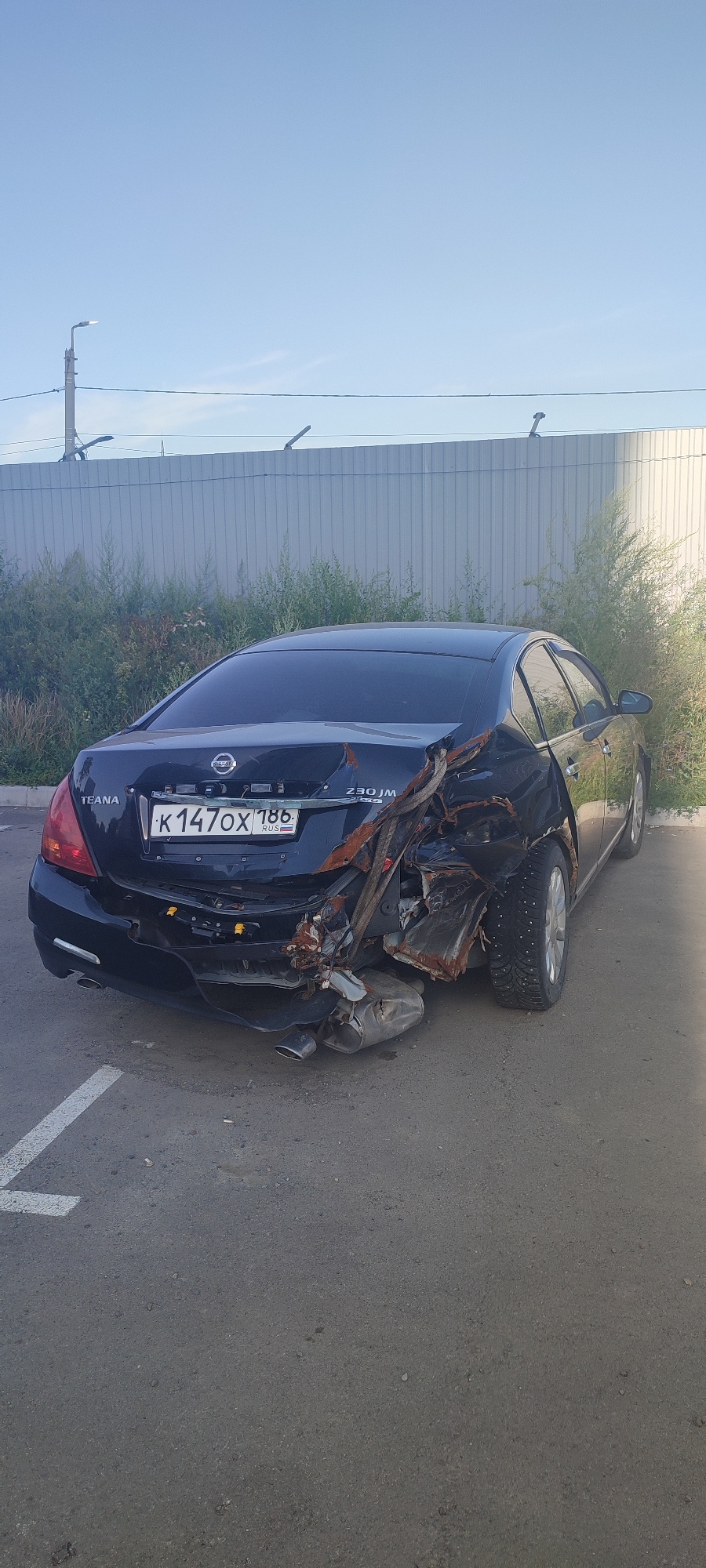 A black sedan, identified as a Nissan Teana with the license plate K147OX 186, is parked on an asphalt surface, likely a parking lot, in Novoaltaysk, Russia. The rear passenger side of the vehicle has sustained significant damage, with the body panels crumpled and twisted, exposing rusted metal components and structural damage. A section of the exhaust system is visible hanging low on the rear of the car. The car is parked next to a tall, corrugated metal fence, with sparse vegetation growing behind it. A street lamp is visible in the distance. The sky is clear and blue. The scene appears to be an outdoor urban environment, possibly on a sunny day.