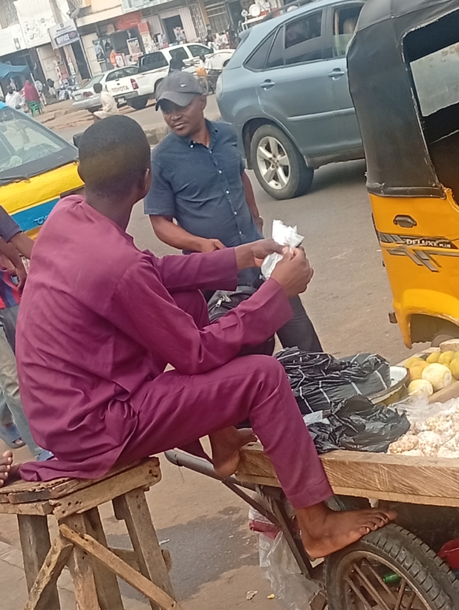 In Maiburiji, Nigeria, a man in a maroon outfit sits on a worn wooden stool next to a hand-pulled cart on a city street. The cart displays bags, lemons, and some unknown produce. A man in a blue shirt and baseball cap stands facing the seated man, who holds a small clear bag in his hands. A three-wheeled yellow "Keke Napep" tricycle is positioned to the right of the men, partially blocking the view. In the background, other cars, buildings, and pedestrians are visible along the street.