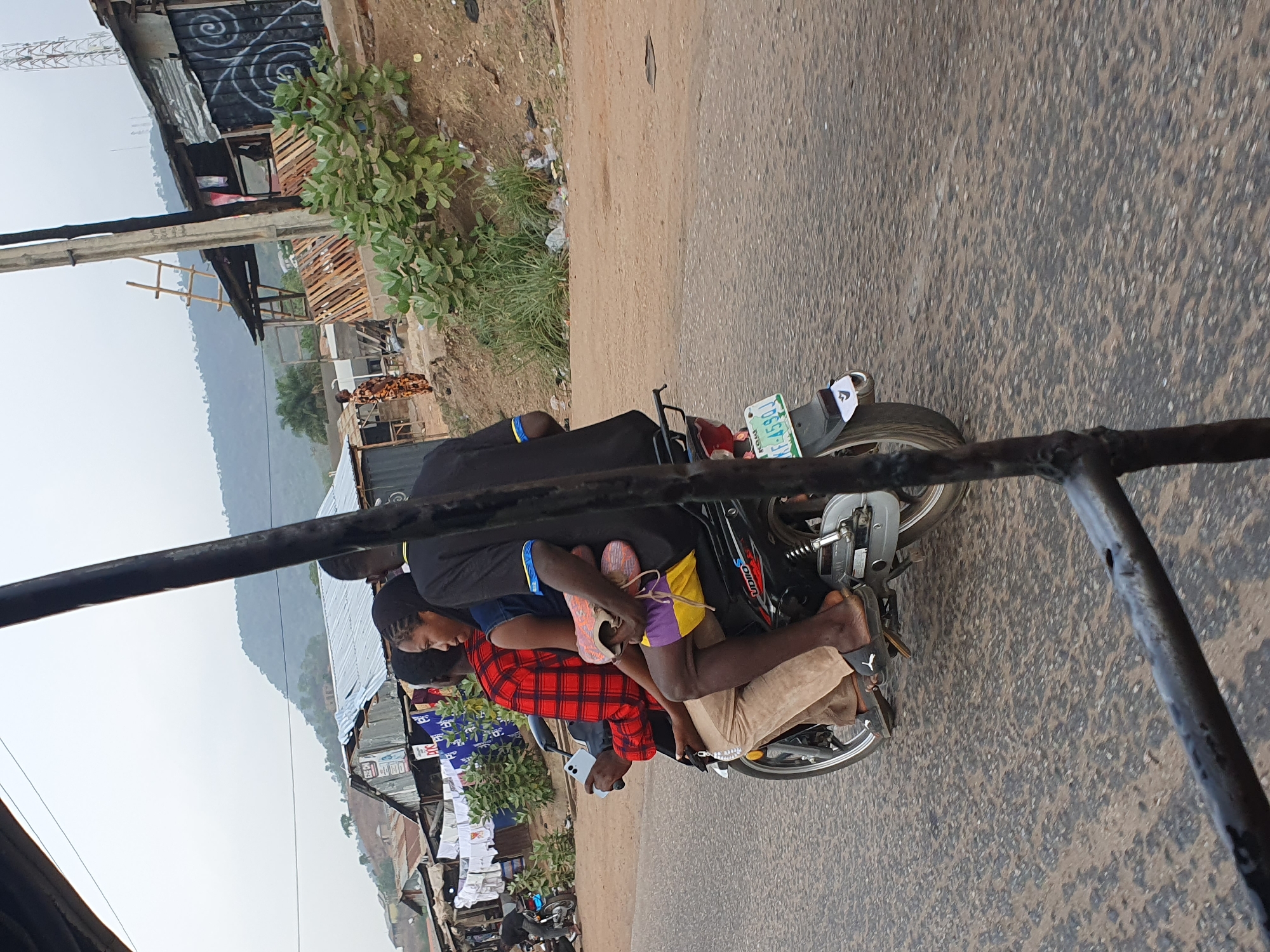 The image, taken in Lokoja, Nigeria, captures a dynamic street scene featuring multiple individuals riding a single motorcycle. The photograph is oriented sideways, but depicts a road stretching into the distance with makeshift structures and hills in the background.
On the black "VIDIO" brand motorcycle, there are at least four people. The driver, mostly obscured by the frame and wearing a black top, is at the front. Immediately behind the driver is a person wearing a red and black plaid shirt, holding a white smartphone. Behind them, a younger individual or child is seated, dressed in a blue shirt and distinct yellow and purple shorts, holding a pair of sneakers. Another leg is visible further back, suggesting at least one more person is also on the overloaded motorcycle. The riders appear calm, focused on the journey.
The motorcycle displays a green and white Nigerian license plate with the registration "KE 459DJ" and the state name "KOGI." The road itself is asphalt, showing signs of wear. Along the roadside, various informal buildings or stalls are constructed from corrugated metal sheets and wooden pallets. One building features distinct white spiral graffiti on a dark wall. Green vegetation, including bushes and small trees, lines the road and surroundings. In the background, rolling hills or mountains are visible under an overcast sky, suggesting either early morning, late afternoon, or cloudy weather conditions. The scene reflects a common mode of transport and typical roadside environment in many Nigerian urban or semi-urban areas.