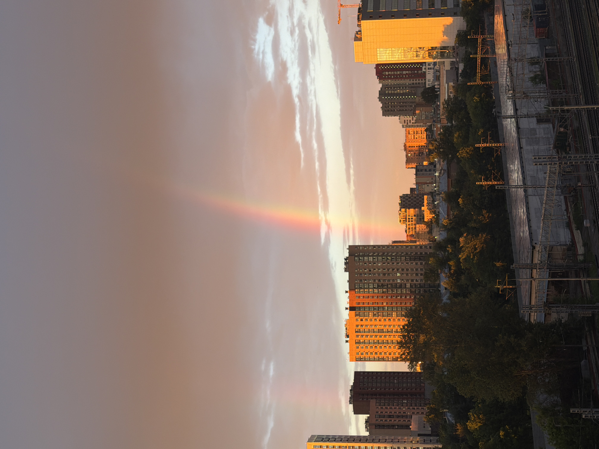This media file captures a sunset over a cityscape in Moscow, Russia. The scene is dominated by towering residential buildings, some of which are bathed in the warm, golden light of the setting sun. The sky is a dramatic canvas of soft oranges, pinks, and yellows, with streaks of white clouds adding texture. A faint rainbow arcs across the sky, a beautiful atmospheric phenomenon often seen during or after rain when the sun is low.
Adjacent to the buildings, a dense cluster of green trees creates a contrast between the urban architecture and nature. Further to the right, a complex network of railway tracks and overhead power lines suggests a transportation hub or industrial area within the city. The overall mood is serene and picturesque, conveying the beauty of a city at dusk. The time of day is clearly evening, given the low angle of the sun and the warm lighting. No people are visible in the frame, and no text is discernible.