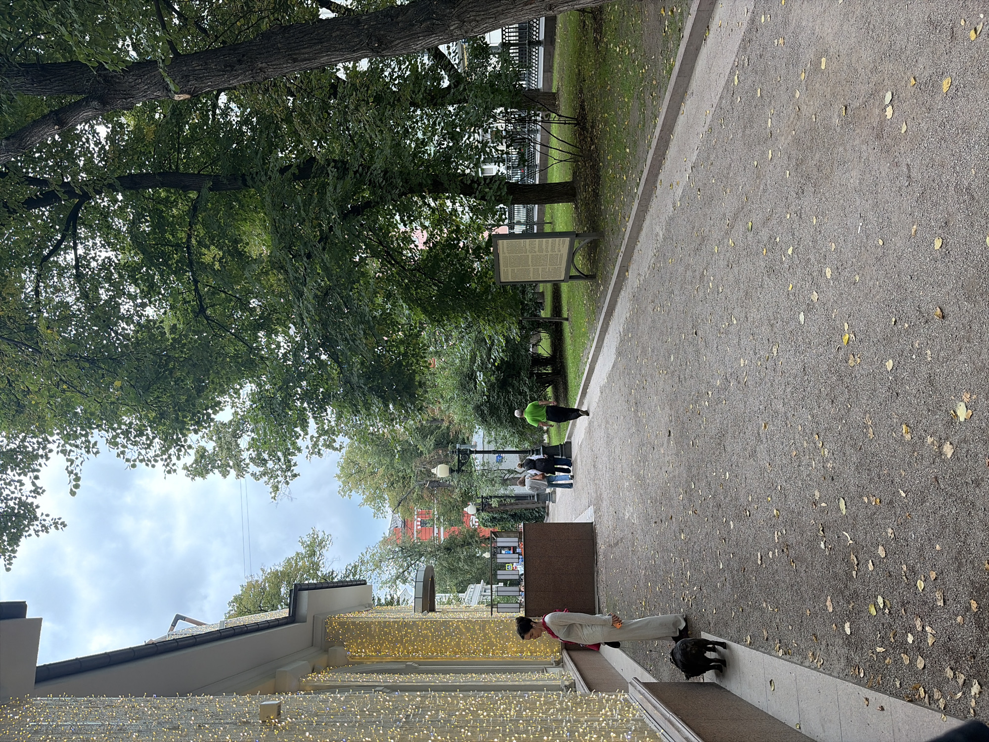 The image depicts a street scene in Moscow, Russia, with a building adorned with numerous small, warm-toned lights on its facade. A person, dressed in light-colored clothing and a pink scarf, stands near the building, accompanied by a black dog. To the right of the building, a pathway made of gravel stretches into the distance, bordered by trees and greenery. Several other individuals are visible walking along the pathway, one wearing a bright green shirt. In the background, more trees and buildings are present, with a lamppost and an information board positioned along the path. The ground is covered with fallen leaves, suggesting an autumn setting. The sky is overcast.