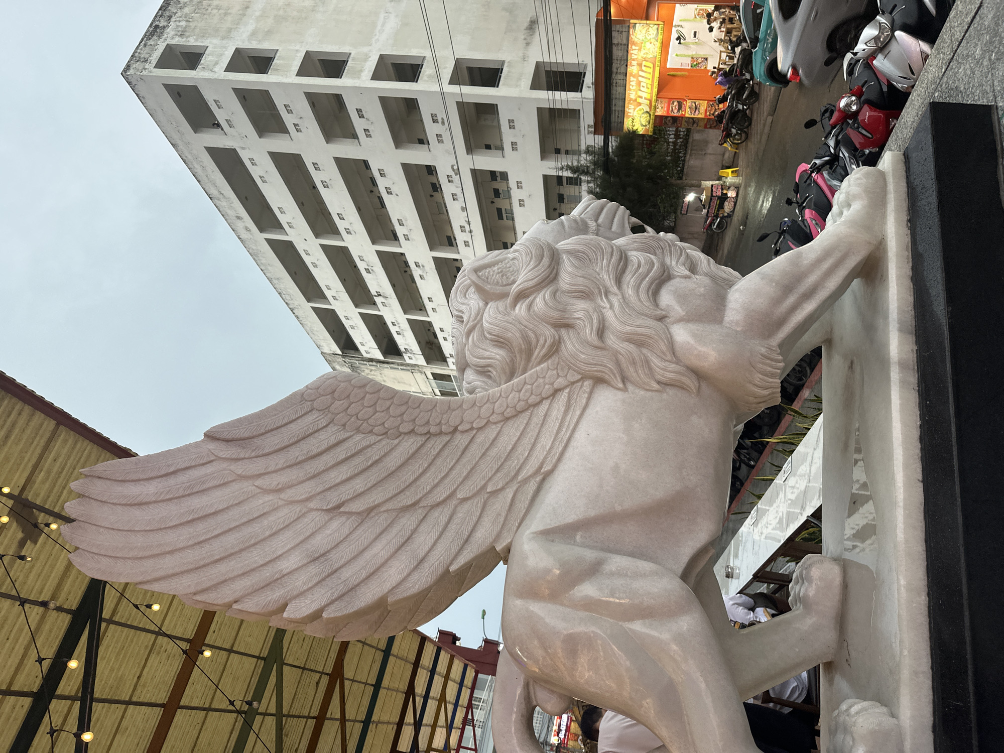 A white marble winged lion statue is prominently featured in the image. The statue stands adjacent to a vertical white marble slab and a dark vertical slab. Motorbikes are parked nearby along the street. Behind the statue is an unfinished, tall building with a repetitive pattern of square openings. Additionally, there is a sign visible that says "HELLO" with Vietnamese characters. The scene is located in the city of Bac Nha Trang Ward, Vietnam. The overall light suggests an overcast day or dusk.