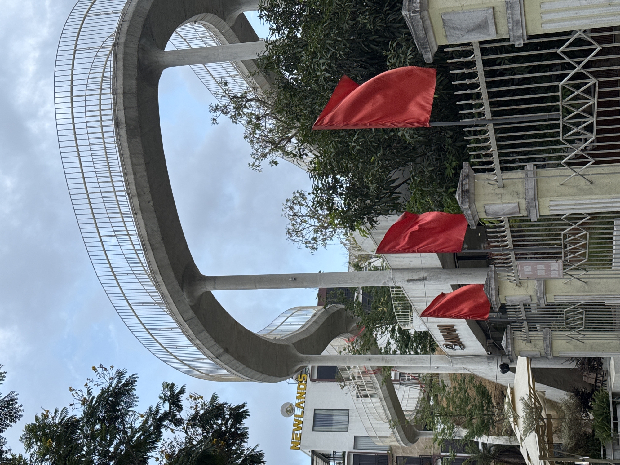 The image shows a street scene in Đà Nẵng, Vietnam, featuring a pedestrian overpass that is composed of a curved concrete structure with metal railing. The overpass spans a street lined with buildings, trees, and flags. A building with the sign "NEWLANDS" is visible. Red flags are mounted on poles adjacent to a fence. The sky is overcast.