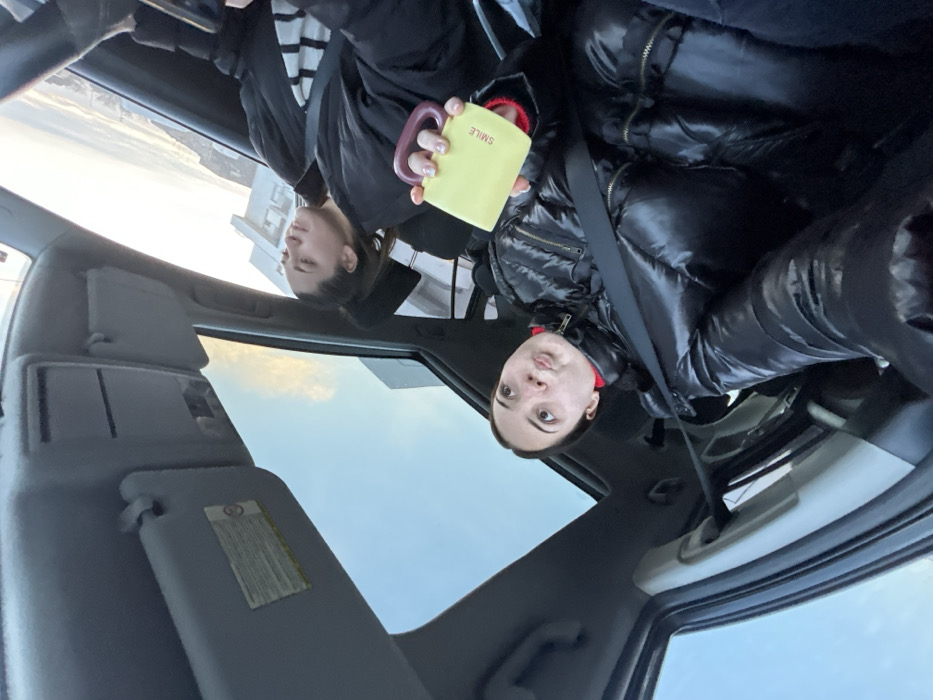 The image captures a casual scene inside a car, seemingly during the daytime in Hoenheim, France. The primary subject is a young woman, seated on the right side of the car, wearing a shiny black puffer jacket over a red garment, with a seatbelt visible across her shoulder. She holds a light yellow mug with a purple handle, on which the word "SMILE" is clearly legible. Her expression is neutral or slightly curious as she looks towards the camera.
In the upper-left portion of the image, the reflection of another young woman is visible, likely in an overhead car window or sunroof. She is wearing a black and white striped top under a black jacket and appears to be looking away. The car's interior features prominently, including parts of the headliner, sun visors (one displaying a warning label with small, indistinct text), and a side window.
Through the car's windows and reflections, a bright, clear to partly cloudy sky is visible, along with a distant building, indicating an outdoor, daytime setting. The overall mood is calm, depicting a paused moment, perhaps during a drive or while parked.