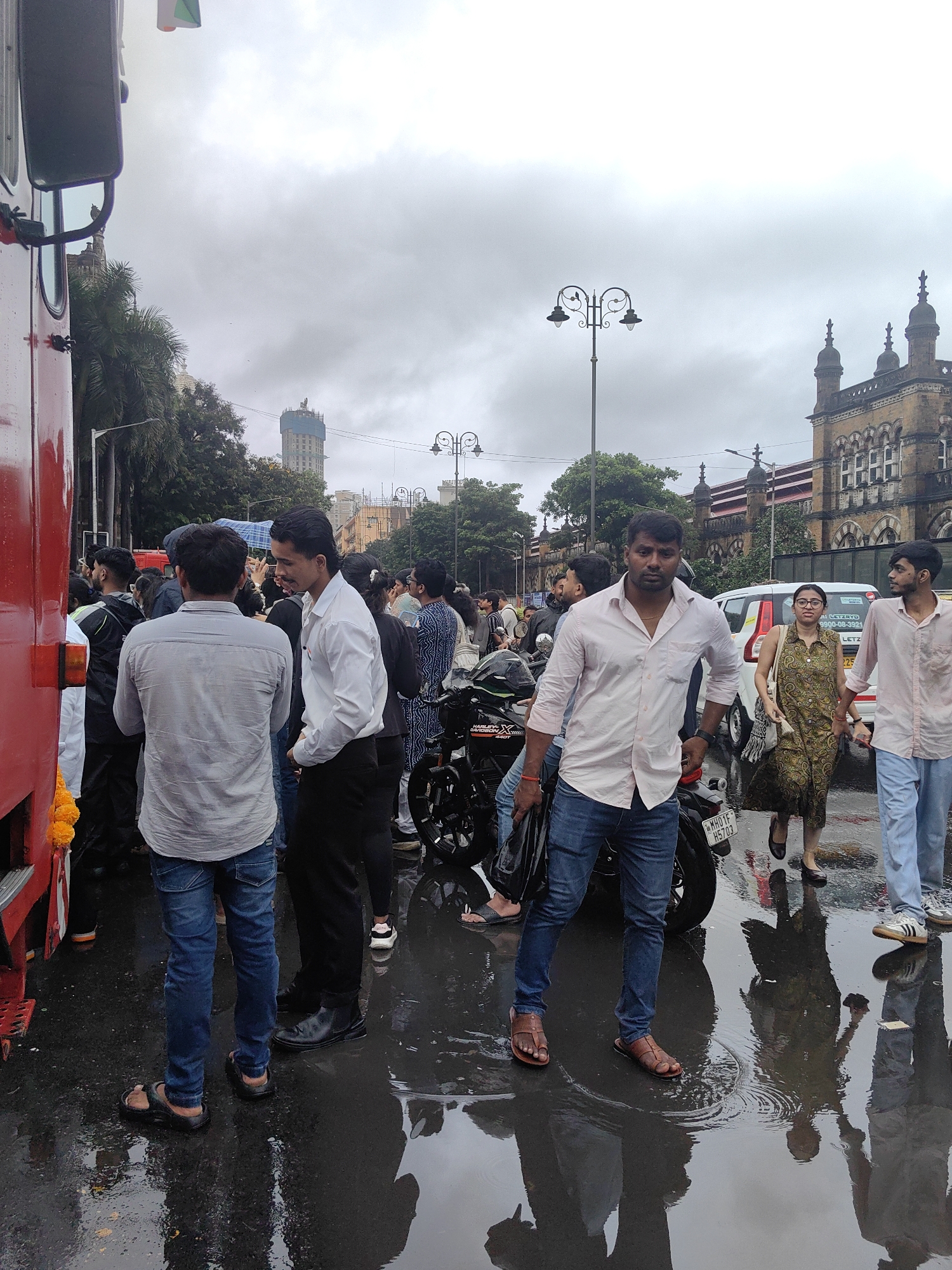 A group of people are gathered on a wet street in front of a red vehicle, possibly a bus or truck. The ground is covered in puddles, reflecting the overcast sky and the surrounding buildings. In the background, architectural structures suggestive of colonial-era design, characteristic of Mumbai, India, are visible, including a clock tower. Some individuals are looking at their phones, while others are engaged in conversation. A motorcycle is parked to the right of the red vehicle. The overall atmosphere appears to be one of an outdoor event or gathering, possibly in a public space in Mumbai.