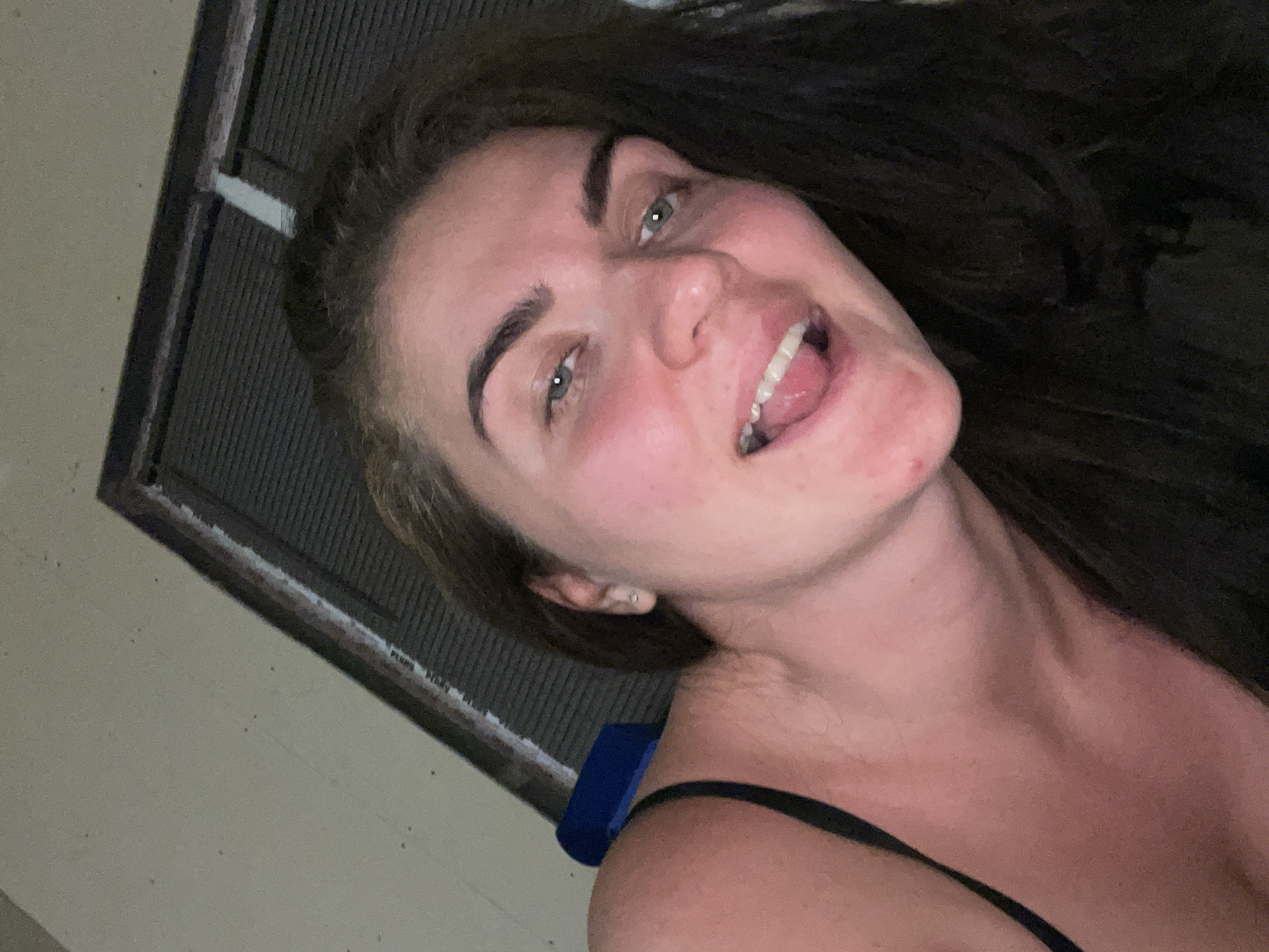 The image shows a close-up selfie of a woman with long, dark hair and green eyes. She is wearing a black tank top and her tongue is sticking out. The background appears to be an outdoor setting with a textured wall and a dark, slatted structure. The lighting suggests it is either dusk or nighttime. No specific location in Мосметаллоконструкция Онт, Russia can be identified from the image.