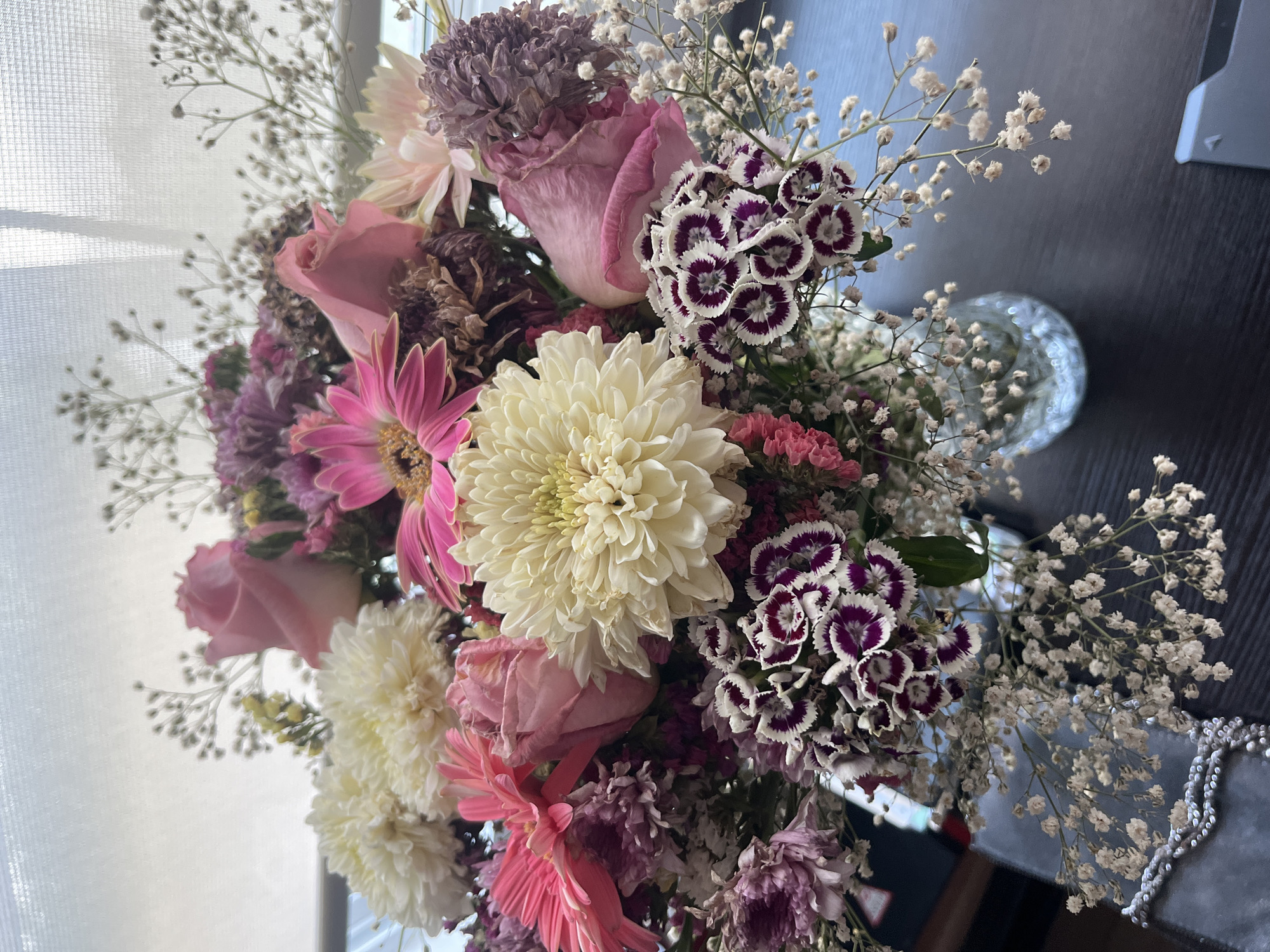 The image shows a close-up of a flower bouquet, featuring a variety of blooms including pink roses, white chrysanthemums, pink gerbera daisies, purple carnations with white edges, and baby's breath. The arrangement is dense and colorful. A vase containing the flowers is partially visible, and a window with sheer curtains is present in the background. The surface on which the bouquet sits is dark and appears to be wood. The image does not provide sufficient information to confirm the location in the city of Barrio de Nuevo León, Mexico, and does not depict any people, actions, or interactions.