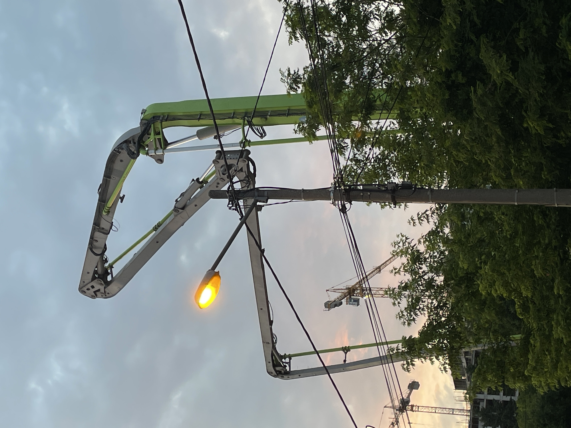 The image shows a concrete pump boom extending upwards against a cloudy sky in Belgrade, Serbia. A street light illuminates the scene, positioned near the boom. Power lines crisscross the view. In the background, construction cranes stand near a building, partially obscured by trees.