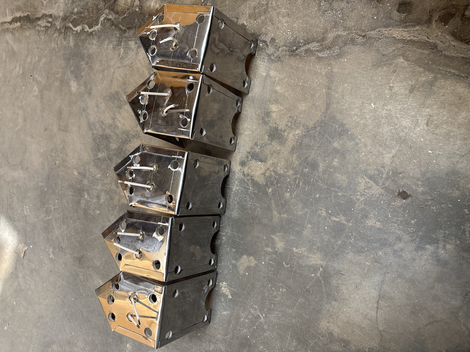 A group of five identical, shiny, metallic objects, resembling small houses with pointed roofs, are lined up diagonally on a grey, concrete surface. Each object has several circular holes on its top surface and two visible holes on its front, lower section. White, string-like material is threaded through the holes on the top of each object. The concrete floor is worn and shows numerous scratches and some patches of discoloration. This scene is located in Telagamurni, Indonesia.