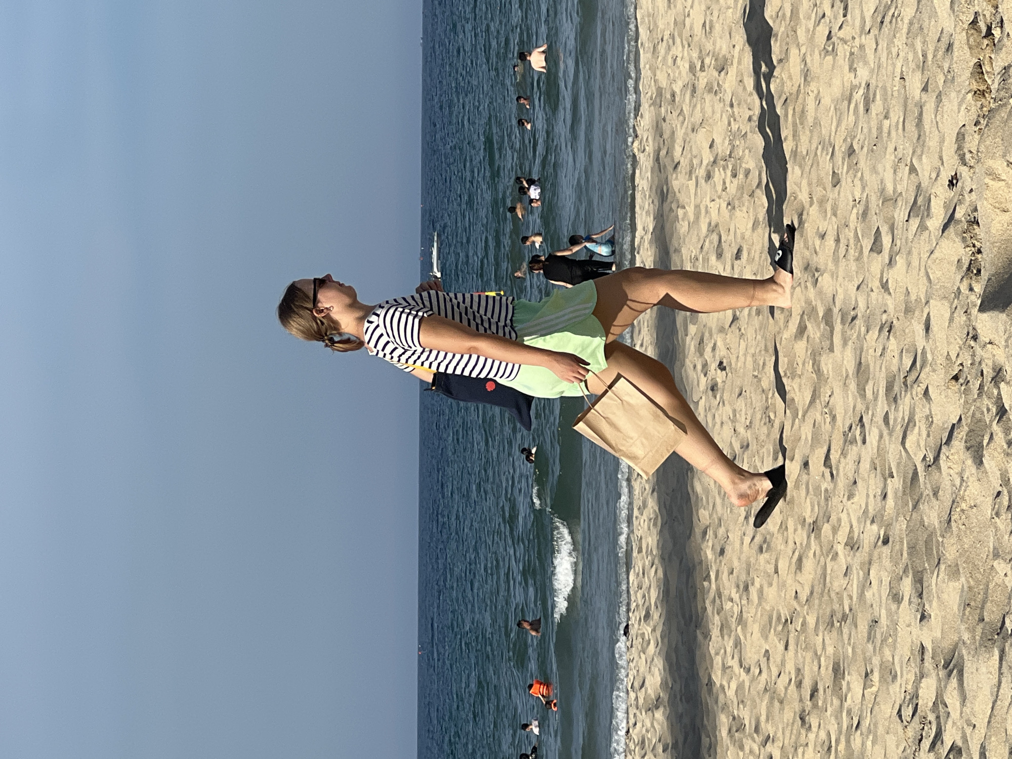 A young woman walks along the sandy beach of Mỹ Khê in Đà Nẵng, Vietnam, carrying a brown paper bag. She is wearing a striped t-shirt, light green shorts, black sunglasses, and black flip-flops. The sea is calm with gentle waves, and several people are swimming and playing in the water. The sky is clear and blue, indicating a sunny day.