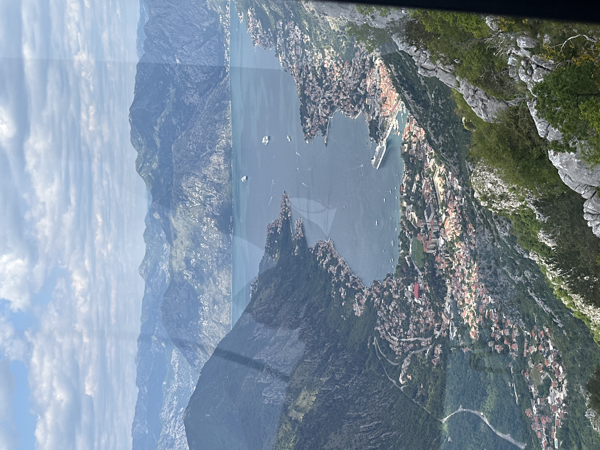 The scene is an aerial view of the city of Škaljari, Montenegro, located on the Bay of Kotor. The bay is visible in the center of the image, with several boats visible. On the right side of the bay and further up the mountain slopes, numerous buildings with red-tiled roofs and various other structures populate the landscape. Jagged, rocky terrain with sparse vegetation is visible on the right side of the frame. On the left, rugged mountains stretch into the distance. The sky above is filled with clouds. The image appears to have been taken from within a vehicle, possibly a helicopter, due to the presence of a dark frame-like border at the top.
