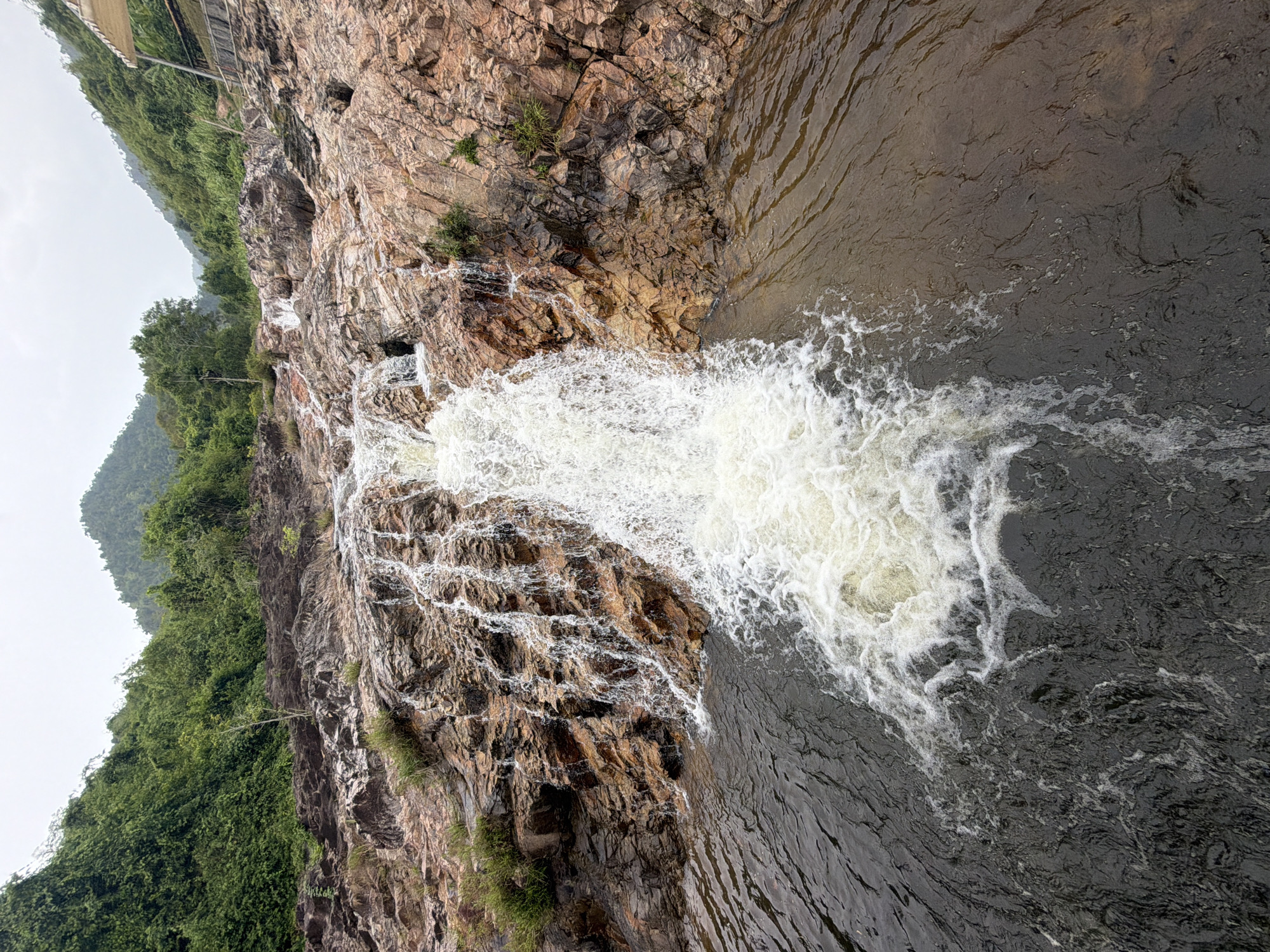 A wide-angle, eye-level shot captures a waterfall cascading over rocky terrain at a location in the city of Khanh Vinh, Vietnam. The waterfall is the primary subject, with white, foamy water tumbling down a series of layered, reddish-brown rocks. The rocks are uneven and textured, with some areas showing moss or small green plants growing from crevices.
The water at the base of the waterfall is dark and appears turbulent, with white foam swirling on the surface. The surrounding environment is lush and green, with dense vegetation covering the hillsides that rise steeply on either side of the waterfall. The trees are a deep green, and the foliage is thick, suggesting a tropical or subtropical climate.
In the background, the sky is overcast, with a soft, diffused light. In the upper right corner of the image, a portion of a simple, open-sided shelter or canopy structure is visible, supported by metal poles and with a light-colored roof. This structure suggests that the area is a tourist or recreational spot.
The overall scene is one of natural beauty, with the powerful force of the waterfall contrasting with the serene green landscape. The image appears to be taken on a day with no direct sunlight.