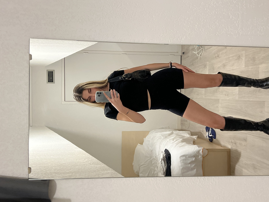 A young woman is captured in a full-length mirror selfie, positioned in an indoor room, likely a hotel room or apartment in Val-d'Isère, France. She has long, straight blonde hair with darker roots and is dressed in a fitted black short-sleeved top, black cycling shorts, and black knee-high boots. A black shoulder bag with a distinct shape is worn across her body. She holds a light grey smartphone with a multi-lens camera in her right hand (left in the reflection), using it to take the photo, while her left hand rests on her hip or thigh. Her expression is neutral, focused on the phone.
The reflected background shows light-colored walls and a light-toned floor, possibly wood or laminate. A bed with white bedding and a light brown headboard or frame is visible in the lower right portion of the reflection. A rectangular vent is present on the wall in the upper left. A pair of blue and white flip-flops are seen on the floor near the bed. The interior appears modern and well-lit, suggesting daytime or ample artificial lighting. No visible text is discernible in the image.