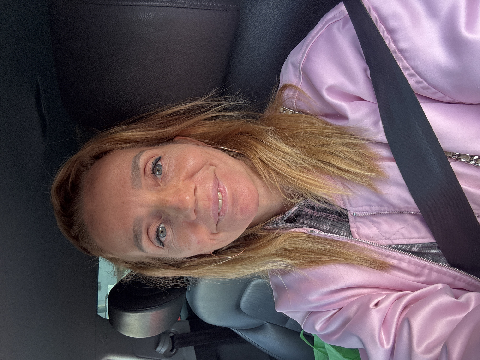 A woman with blonde hair and blue eyes is seated in the driver's seat of a car, facing the camera. She is wearing a pink satin bomber jacket over a plaid shirt and a black seatbelt is visible across her chest. The interior of the car, including a black leather seat and a portion of the dashboard, is visible in the background. The lighting suggests it is daytime, possibly outdoors. The location is not specified as being in the city of Unknown, Russia.