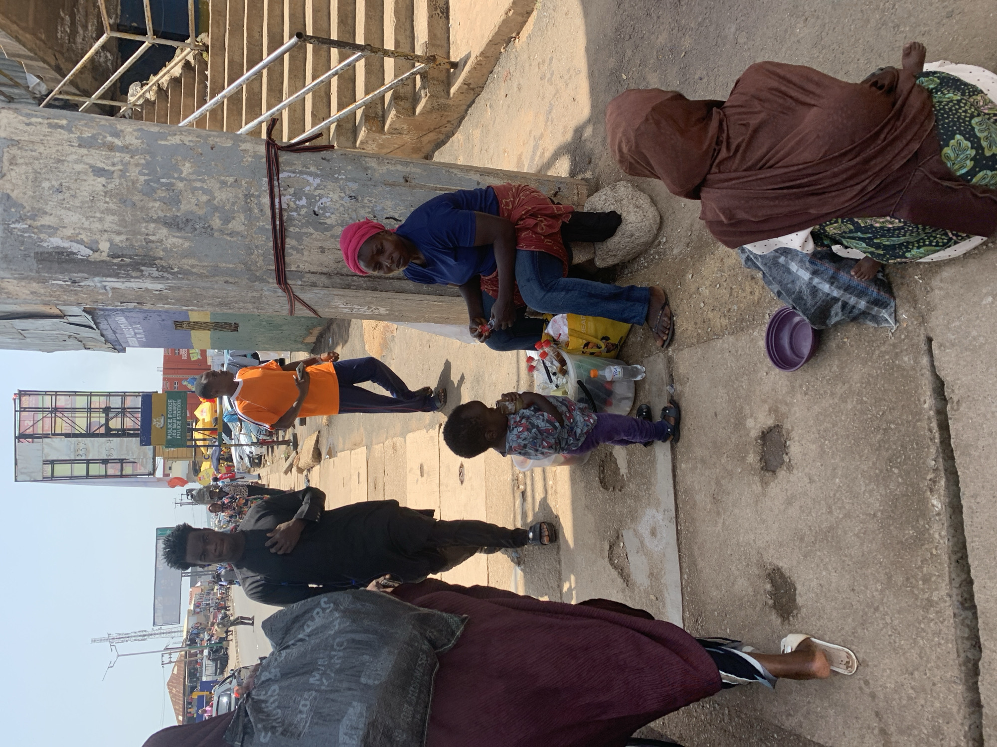 The image depicts a street scene in Jos, Nigeria. In the foreground, a young child wearing a patterned shirt and purple pants stands near a woman in a blue t-shirt and jeans, who is seated on a rock by a concrete pillar. The woman, who is wearing a pink headscarf, is holding a small object and appears to be engaged in a transaction. A yellow shopping bag sits beside her, filled with items, including several bottles of water and a plastic bottle. Behind them, a man in a black outfit walks away from the camera, while another man in an orange t-shirt and blue pants walks towards the right of the frame. A woman in a maroon head covering and dark clothing is partially visible on the left, carrying a large bag. In the background, there are buildings, a street with vehicles, and a tall billboard advertising "FIRST FEDERAL SAVINGS BANK PLC" and "POLICE FORCE JOS". Another billboard with the text "332 854" is also visible. A set of concrete stairs with metal railings is situated to the right of the concrete pillar. The ground is paved with concrete, and there are some potholes and debris. The overall atmosphere appears to be that of a busy urban environment.