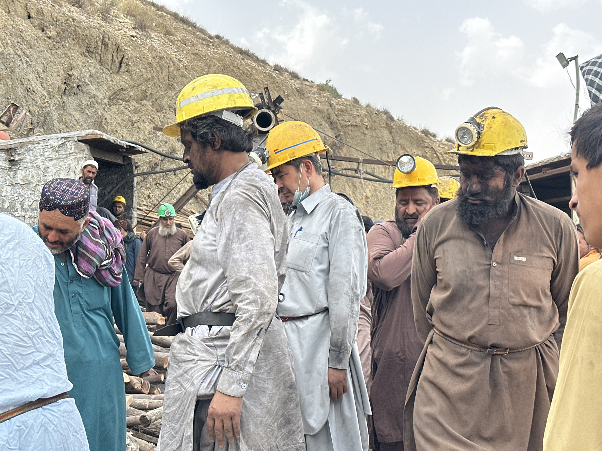 This is an eye-level, medium shot photograph taken outdoors in the city of Quetta, Pakistan, likely near a mountainous or hilly area given the rocky, barren slope dominating the background. The scene appears to be a gathering of men, many of whom are wearing mining helmets. The sky is overcast.
In the foreground, several men are visible. The man on the far left is wearing a light blue garment and has a colorful scarf draped over his shoulders and head. To his right is a man in a teal-colored shalwar kameez with a patterned skullcap. He has a beard and is looking down. Next to him is a man wearing a light gray outfit and a yellow hard hat with a headlamp attached. His face is dirty, suggesting he has been working in a dusty environment. He is looking forward and slightly down. To his right, another man in a gray outfit and a yellow hard hat with a headlamp is also visible, his face obscured by shadow. Further to the right is a man wearing a brown shalwar kameez and a yellow hard hat with a headlamp. His face is covered in dark dust and dirt. He is looking forward and slightly down.
Behind these individuals, other men are visible, some wearing similar clothing and some in hard hats. There is a small, simple structure with a roof to the left of the frame, and piles of cut logs are visible on the ground. The overall atmosphere suggests a somber or serious occasion, possibly related to mining work or an incident. The presence of headlamps on the helmets indicates that the work may involve underground mining.