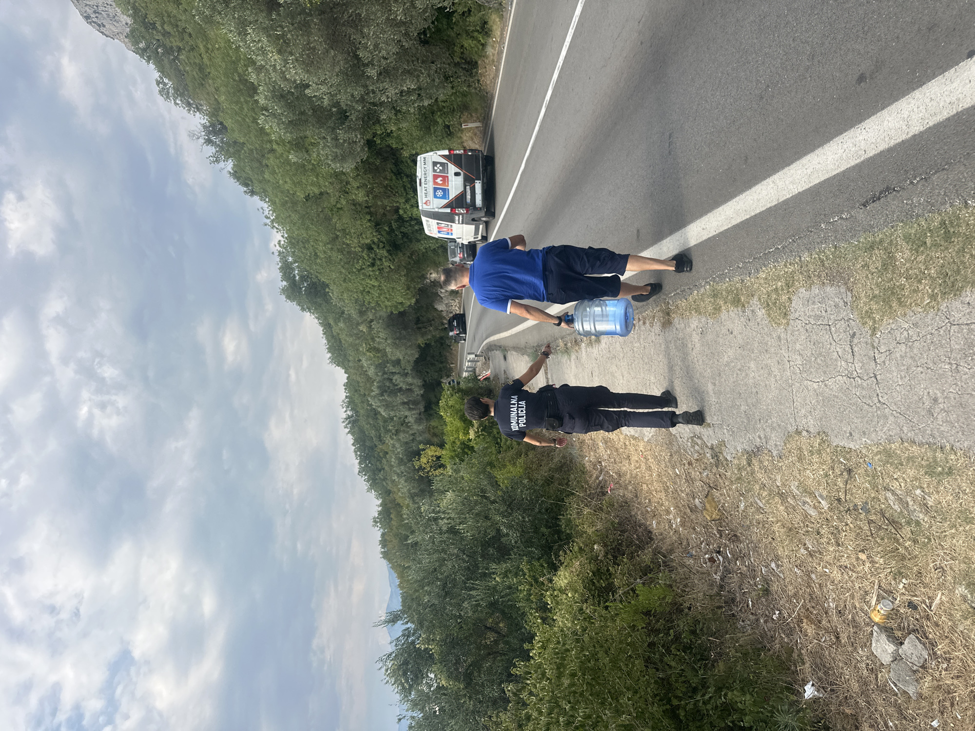 A person in a dark blue uniform with "KOMUNALNA POLICIJA" text on the back walks along a paved path on the side of a road. To the right of the path, a man in a blue polo shirt and dark shorts walks on the road, carrying a large blue water jug. He is walking away from the viewer and towards a white van with "HEAT ENERGY MM" visible on its side. The van is driving on the road. The scene is located in Drobnići, Montenegro, with a hillside covered in dense green foliage to the left of the path and extending up to the right behind the road. The sky is cloudy. The path and the shoulder of the road are littered with trash.