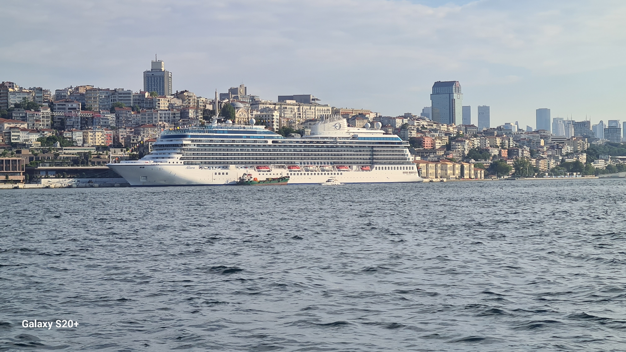 A large white cruise ship, identified by the name "Allure" on its hull, is docked in the foreground of a city skyline. The ship, which takes up a significant portion of the frame horizontally, is positioned on the right side of the image. Behind the ship, a densely populated hillside is covered with numerous buildings of varying sizes and colors, typical of Istanbul's architecture. Modern high-rise buildings are interspersed with older structures, creating a layered urban landscape. A body of water, likely the Bosphorus Strait, occupies the bottom half of the image, with a slightly rippled surface reflecting the sky. A smaller green and white boat is moored alongside the cruise ship, and a white speedboat is seen near the front of the cruise ship. The sky is partly cloudy, with a soft light suggesting it is either early morning or late afternoon. The overall impression is a vibrant city scene with a majestic cruise ship as a focal point.