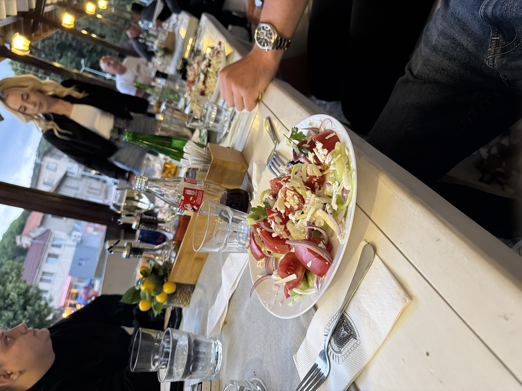 The image depicts a table setting at an outdoor restaurant in Brestovitsa, Bulgaria. A plate of salad, consisting of tomatoes, cucumbers, onions, and feta cheese, is centrally placed. To the left of the salad are two glasses of water, a napkin, and a fork. To the right, a bottle of Coca-Cola and other drinks are visible, along with a wooden container holding cutlery. The background shows people seated at other tables and buildings in the town. The overall atmosphere suggests a casual dining experience.