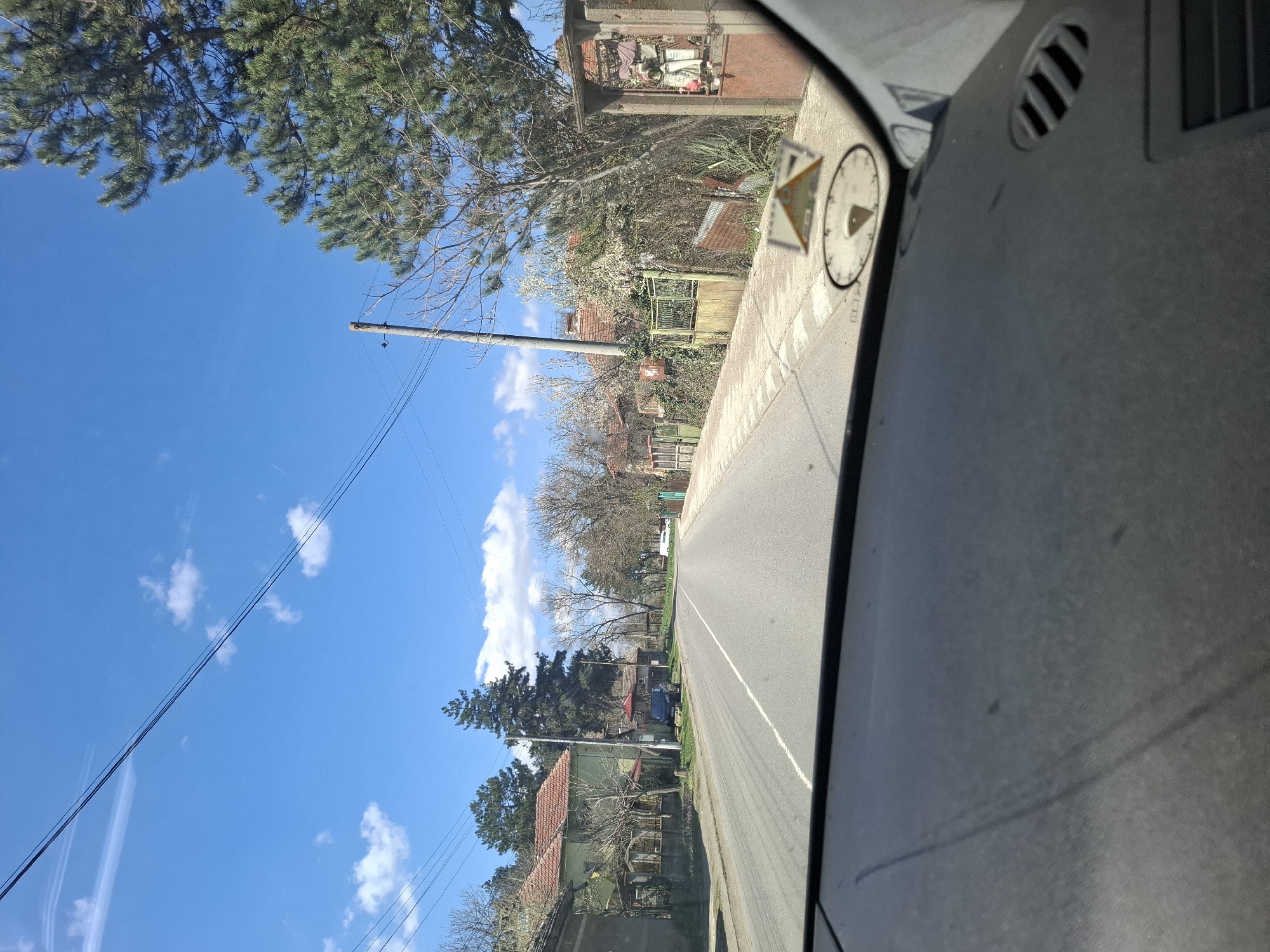 View from inside a vehicle, looking out onto a paved road in what appears to be a residential area. The road has a white line marking the center. Houses with red tiled roofs are visible on both sides of the road, partially obscured by trees and greenery. Utility poles and power lines run alongside the road. In the foreground, part of the vehicle's dashboard is visible, including vents and a small clock or gauge mounted on the dashboard. The sky is blue with scattered clouds. Based on visual characteristics, this is Studeno Buche, Bulgaria.