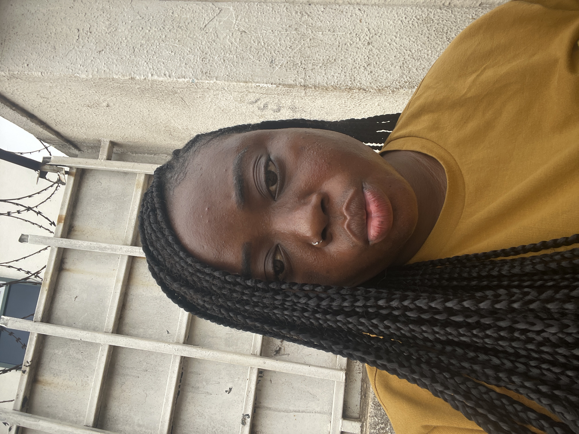 A medium shot shows a Black woman with braided hair, wearing a yellow top, with a nose ring, positioned near a gray metal fence topped with barbed wire. The location is determined to be in the city of Abuja, Nigeria.