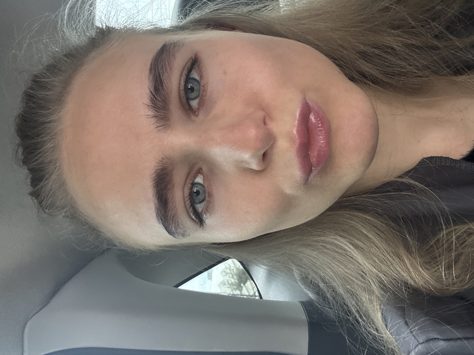 A close-up, slightly angled headshot of a woman with fair skin and blonde hair. She has light blue eyes, prominent eyebrows, and full lips with a glossy sheen. She appears to be inside a vehicle, suggested by the glimpses of the car's interior. The exact location in Unknown, Unknown is not discernible from the image.