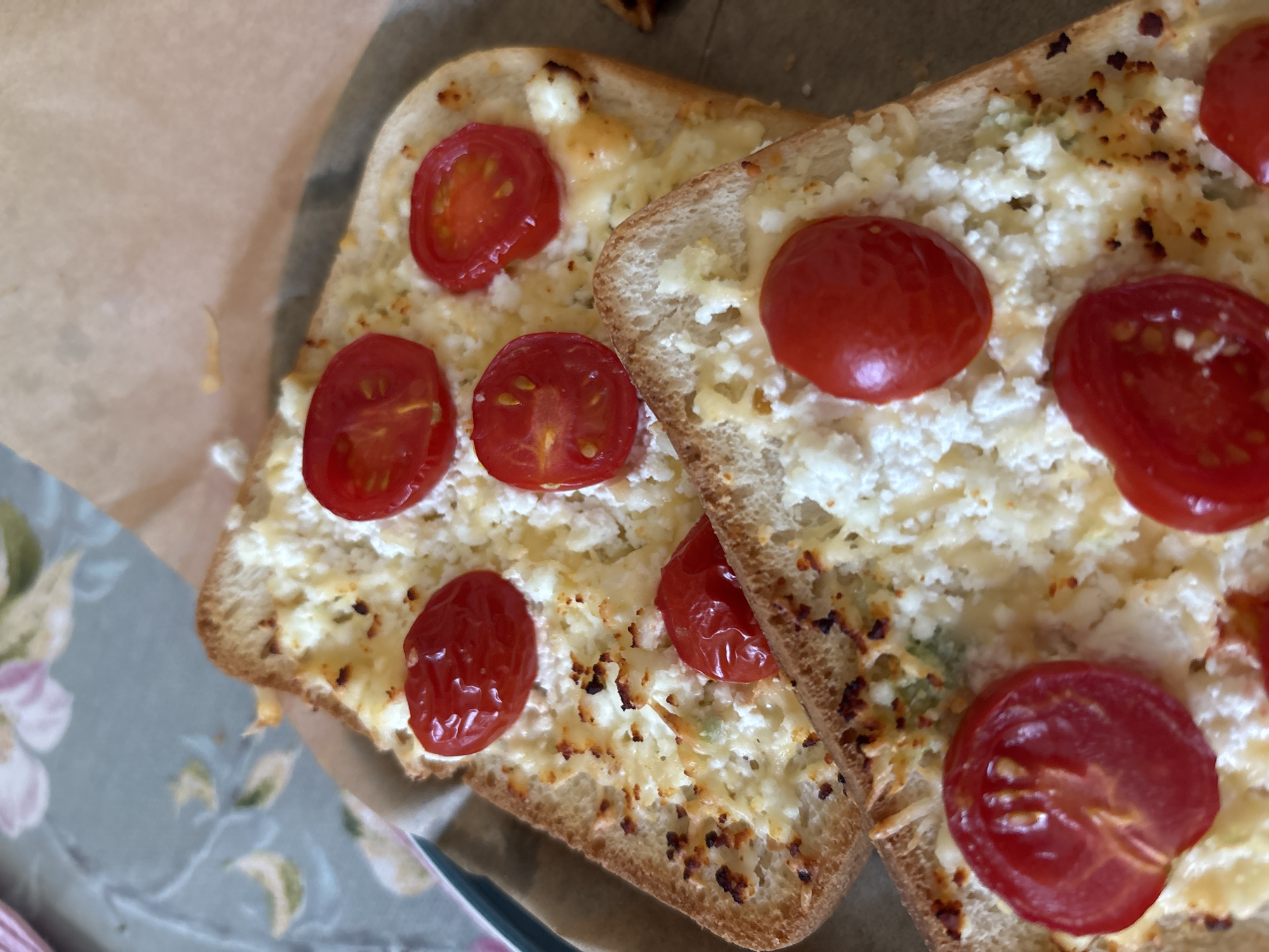 Two slices of toast with cheese and cherry tomatoes are arranged on a brown paper surface, overlaid on a patterned tablecloth. The toast slices are topped with crumbled white cheese and halved cherry tomatoes. The cheese appears slightly melted and browned in places. Some of the cherry tomatoes are whole, while others are cut in half, revealing their inner texture. The background shows a floral patterned tablecloth in muted colors of green, pink, and beige. The image is taken from a top-down perspective. The location is not specified.