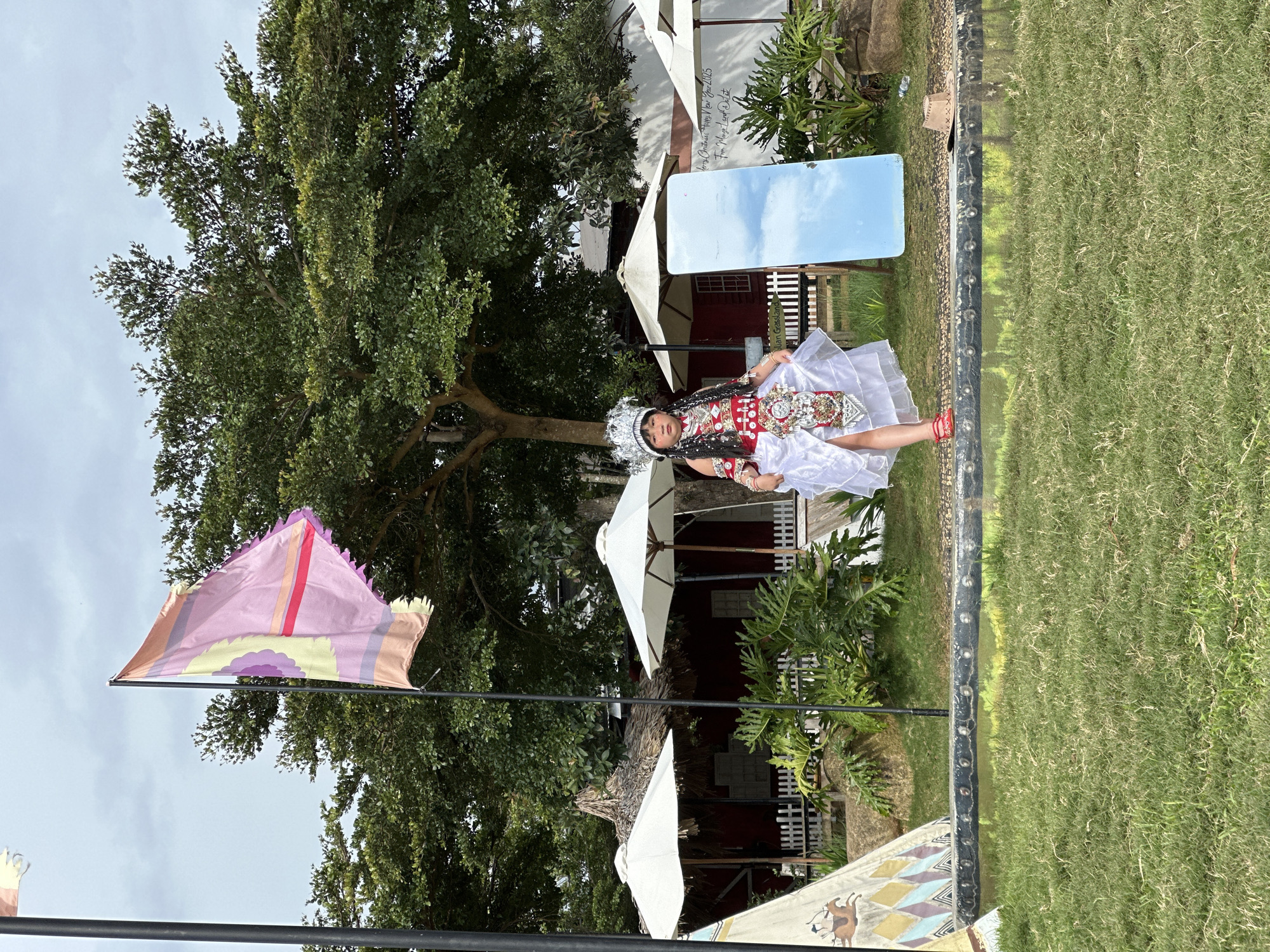 A young girl, dressed in a traditional white and red outfit with an elaborate headdress, stands in front of a red building with thatched-roof awnings and white umbrellas. She is positioned on a raised platform overlooking a grassy area and a reflective pool. Behind her, a large tree provides shade, and a flag with pink and yellow stripes flutters in the wind. To her right, a tall mirror reflects the sky. A cowboy hat lies on the ground near the edge of the platform. The setting appears to be a recreational or cultural space within Thôn 5, Vietnam, characterized by lush greenery and rustic structures.