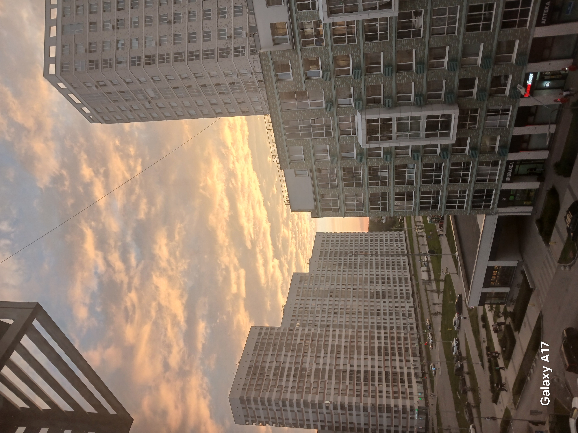 The image shows a view of a residential area at sunset, likely in Yekaterinburg, Russia, given the "Galaxy A17" watermark, a model commonly sold in that region, and the Cyrillic text on storefronts. In the foreground, there is an overhead structure, possibly a balcony or pergola, casting a shadow. The sky is filled with dramatic clouds tinged with orange and yellow from the setting sun.
Dominating the mid-ground and background are several modern, multi-story apartment buildings. The building on the right is tall and slender with a light-colored facade, featuring many uniformly spaced windows. To its left, a much larger, sprawling apartment complex with a white facade and numerous windows stretches across the frame. In the foreground and mid-ground, a lower building with a greenish-gray brick facade and large windows is visible. This building appears to house commercial establishments on the ground floor, indicated by signage in Cyrillic, including "ПИВЛАВКА" and "АПТЕКА".
Below the buildings, a street scene unfolds. Cars are visible on the road, and pedestrians are walking on sidewalks and in a park-like area. Several streetlights are present. The overall impression is a contemporary urban landscape bathed in the warm glow of twilight.
