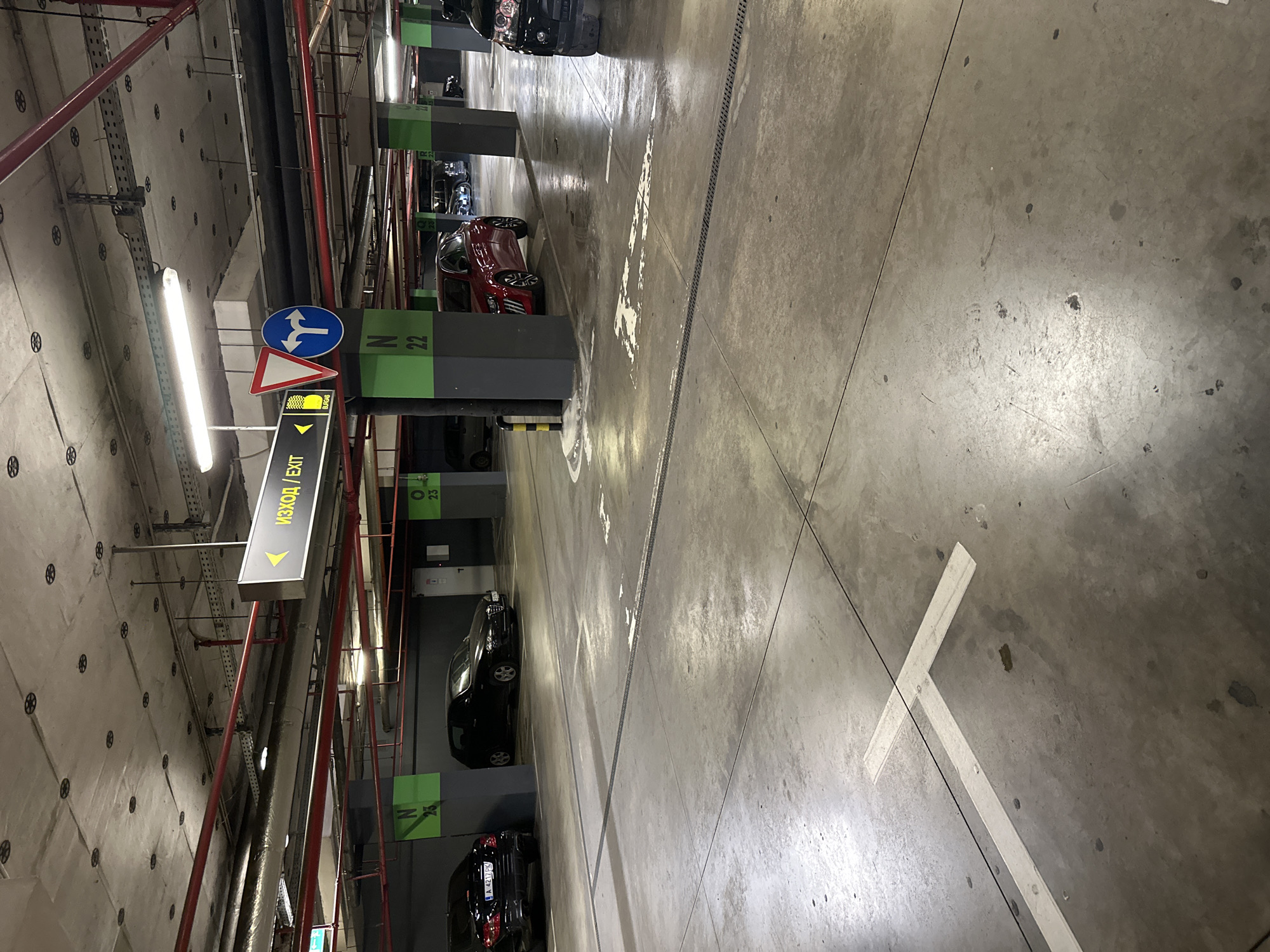 This image captures a wide view of an underground parking garage in Burgas, Bulgaria. The concrete ceiling features exposed infrastructure including red fire suppression pipes, grey insulated ventilation ducts, electrical conduits, and numerous circular black mounts. Fluorescent lighting fixtures are attached to the ceiling, providing artificial illumination.
The floor is grey concrete, marked with faded white lines indicating parking spaces, a "T" intersection, and directional arrows. Black metal drainage channels are embedded into the floor surface. Several dark grey concrete columns with green painted tops are visible, displaying white alphanumeric identifiers such as "N 23," "N 22," and "O 23."
On the far left, a black Mercedes-Benz sedan with Bulgarian license plate A 4217 PK is parked. To its right, a black Alfa Romeo 147 hatchback is also parked. A red Peugeot 208 hatchback is positioned in the middle-right of the frame. The front of a black SUV is partially visible on the far right. Additional vehicles are parked further down the aisle in the background.
An overhead black sign with a yellow left-pointing arrow displays "Изход / Exit" in Cyrillic and English. Below it, a smaller yellow sign reads "БУРГАС" (Burgas). Mounted on a pole to the right of the overhead sign are two road signs: a white triangular "Give Way" sign with a red border and a blue circular "Turn Left or Right" sign. A yellow and black striped bollard is situated next to the column marked "N 22". No people are visible within the scene.