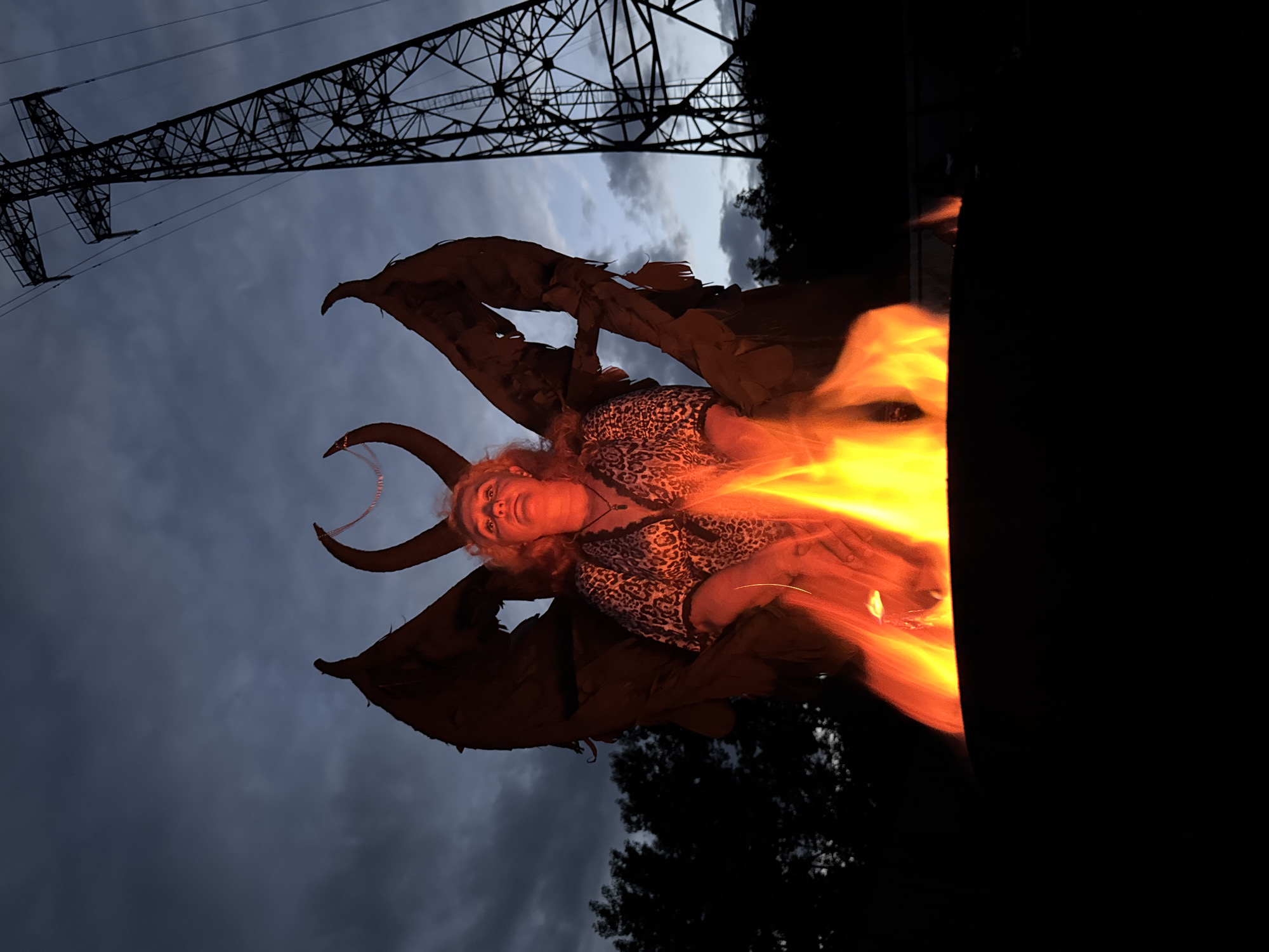A medium shot captures a person sitting in a costume resembling a demonic figure, complete with large, tattered wings and horns. The individual is seated in a fire pit, with flames prominently visible in the foreground. The backdrop features a darkening sky with clouds, trees silhouetted against the horizon, and a large electrical pylon on the right side of the frame. The scene appears to be set outdoors, likely in a rural or industrial area, given the presence of the pylon. The lighting is dim, with the fire providing a strong orange and yellow glow that illuminates the subject and casts shadows. The person is wearing a leopard-print top. The specific location in Chaikovsky, Russia, cannot be determined from the image alone.