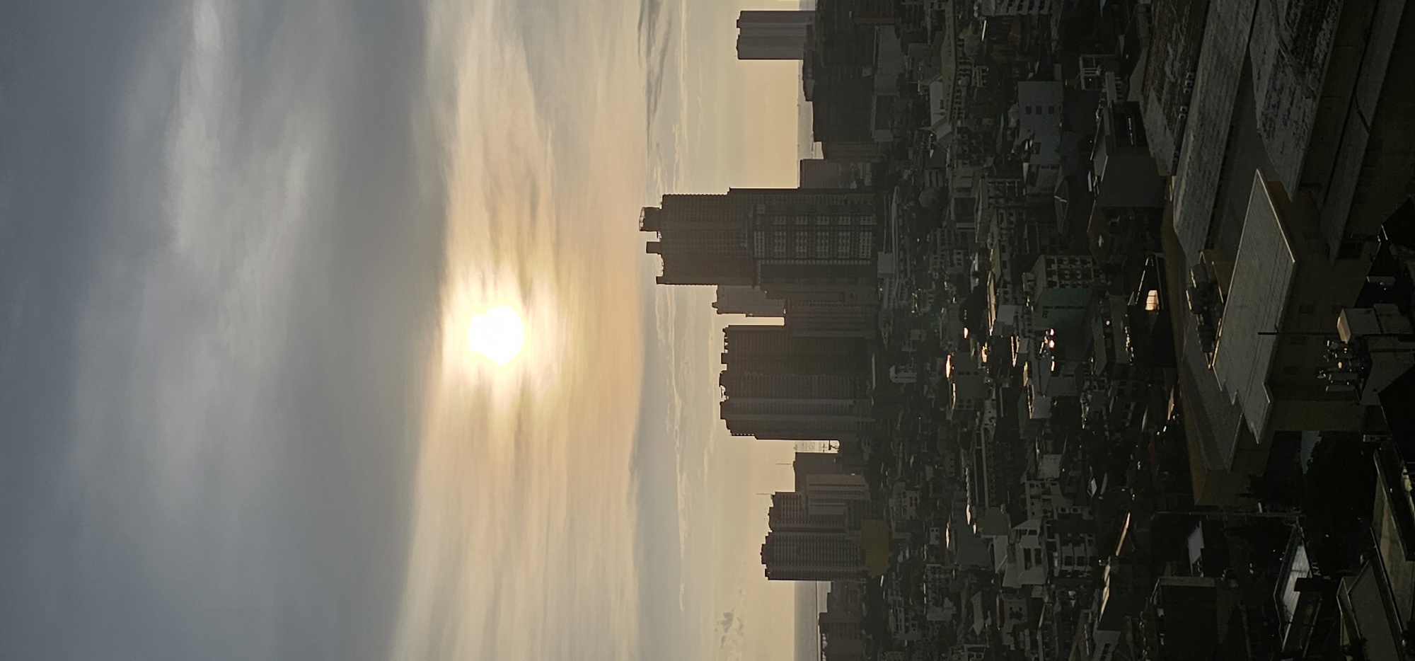 The image is a vertical, aerial photograph of the Makati Central Business District in the Philippines at sunset. The sky is filled with overcast clouds, with the sun appearing as a bright, diffused disk near the center. Below the sky, a dense urban landscape of buildings of varying heights stretches across the frame. In the middle ground, several tall, modern skyscrapers dominate the skyline, some appearing to be under construction. In the foreground and extending into the middle ground, there is a densely packed area of smaller buildings, many with flat roofs. The lighting suggests a low sun angle, casting long shadows and creating a warm, golden hue on some surfaces. The overall impression is one of a bustling, large city at the end of the day, with the natural light interacting with the man-made structures. There are no visible people or specific actions depicted. The environment is purely urban, with a hint of a body of water in the distance on the left side of the frame, likely Manila Bay.