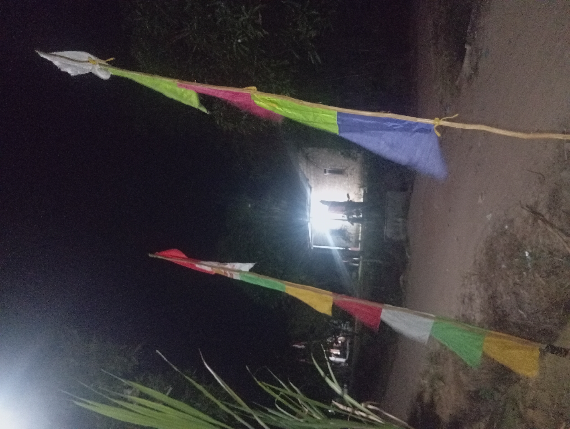 The image, taken at night in Dente Teladas, Indonesia, depicts two colorful flags on poles in the foreground, set against a dimly lit rural landscape. The flag on the left features sections of red, white, green, and yellow. The flag on the right has sections of white, green, pink, and blue. Both flags appear to be made of fabric.
In the background, a simple building with illuminated windows is visible, suggesting a dwelling or small establishment. The surrounding area is characterized by a dirt path and vegetation, including tall green stalks on the far left. The overall atmosphere is dark and somewhat hazy, likely due to artificial lighting and the nighttime conditions. The scene appears to be outdoors, possibly in a village or open area within Dente Teladas. There are no clearly identifiable individuals engaged in any specific actions.