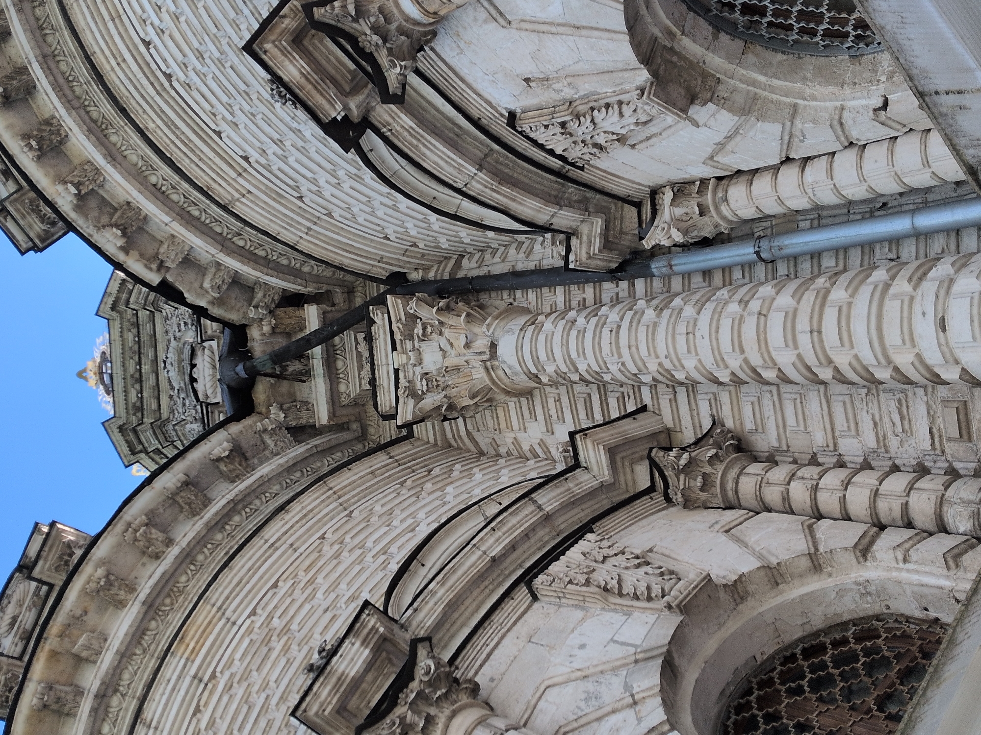 The image shows an upward-looking view of architectural details in Dubrovitsy, Russia, possibly part of the Church of the Sign of the Holy Virgin. The architectural elements include elaborately carved stone arches, columns, and moldings. The sky is visible in the upper left corner. A metal pipe runs along part of the structure. The overall impression is one of ornate, historic construction.