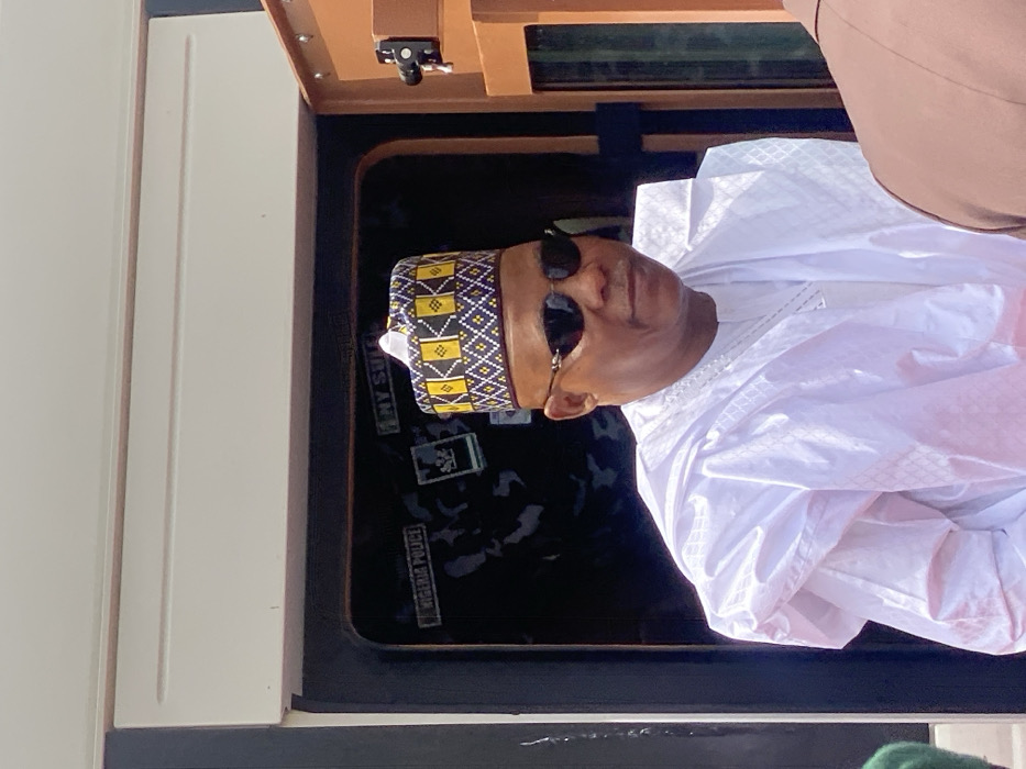 An adult male is prominently featured, wearing a white traditional Nigerian agbada with subtle textured patterns and a distinctive hat (fila) adorned with yellow, black, and white geometric designs. He wears dark, rimless sunglasses and a silver-colored chain with a pendant is visible around his neck. The subject is oriented slightly to his right (viewer's left), maintaining a neutral expression. He appears to be standing or positioned at an entrance.
Behind the subject is a dark opening, likely a vehicle interior or doorway, where a white label clearly displays "NIGERIA POLICE." Above this, another partial label reads "MY SUI." Faint dark grey patterns, possibly camouflage, are visible on the surface behind the labels. To the right of the subject, a brown structural element, consistent with a vehicle door frame, is visible, featuring a black lock mechanism. Above the subject, a light-colored, paneled overhead structure is present. A small portion of green fabric is visible on the far left of the frame, and a reddish-colored surface is visible at the bottom. The scene is well-lit, suggesting outdoor daylight conditions. The location is in Ifira, Nigeria.