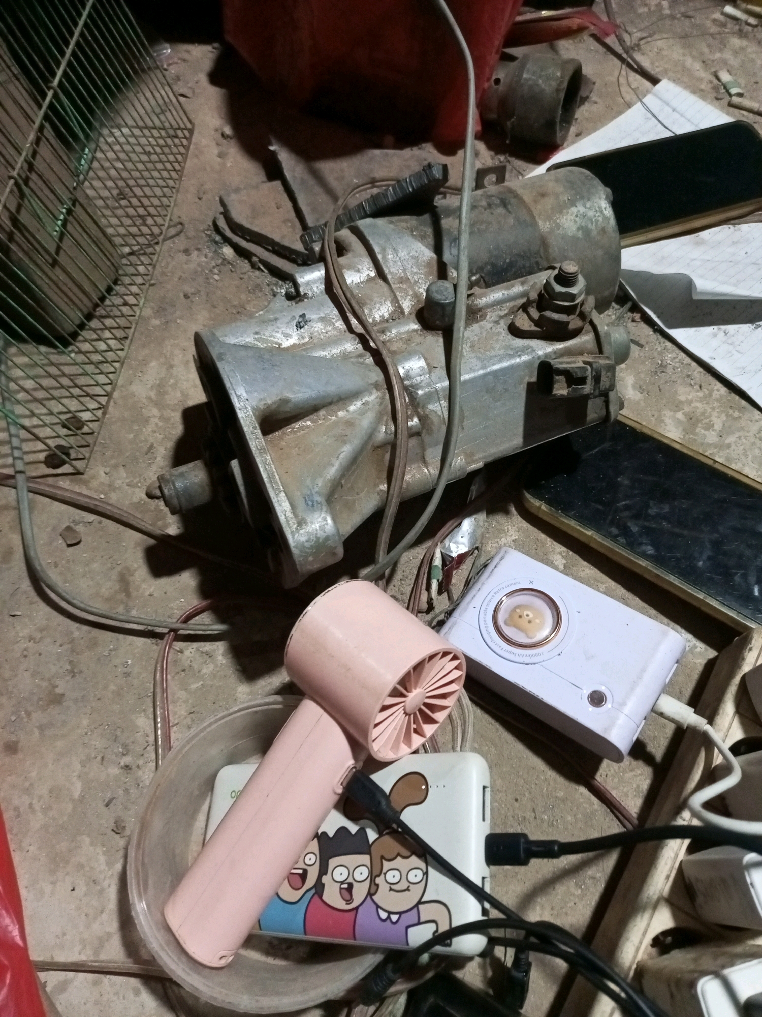 In the city of Keling Panggau, Indonesia, a cluttered, dusty, concrete floor is the setting for an assortment of electronic devices and salvaged parts. A large, metallic starter motor, covered in grime and rust, dominates the center of the image. It is surrounded by various cables, some plugged into power banks, others loose. A small, pink handheld fan rests within a clear plastic bowl, next to a white power bank adorned with cartoon characters. Another white power bank, smaller and circular, sits on top of a black smartphone. The background shows a green metal cage, a reddish-orange object, and more discarded items, all bathed in dim, overhead lighting. The scene appears to be an outdoor workspace or storage area, likely in a residential or informal commercial setting.