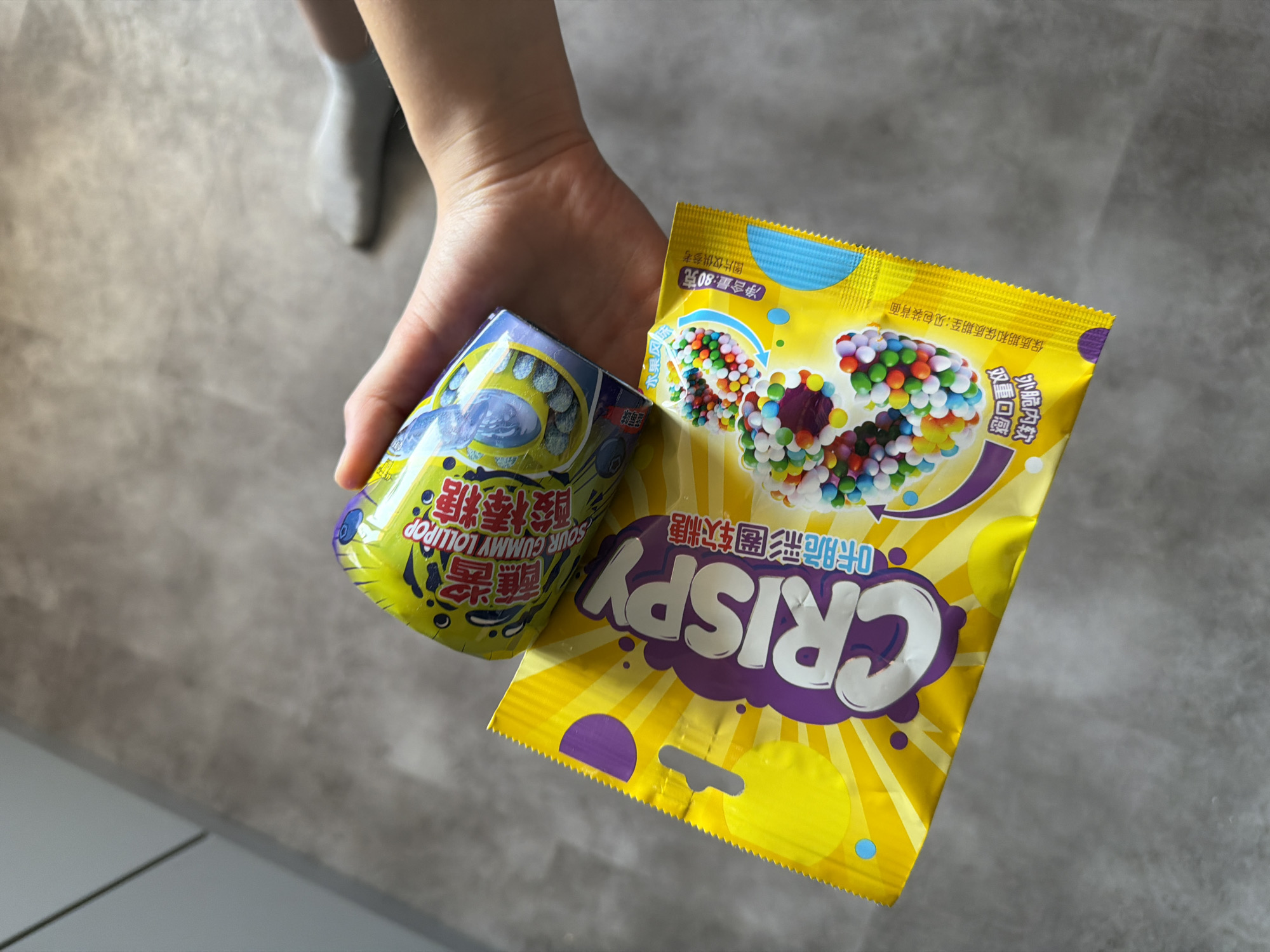 A hand holds a can of "SOUR GUMMY LOLLIPOP" and a yellow package labeled "CRISPY." The location is unknown.