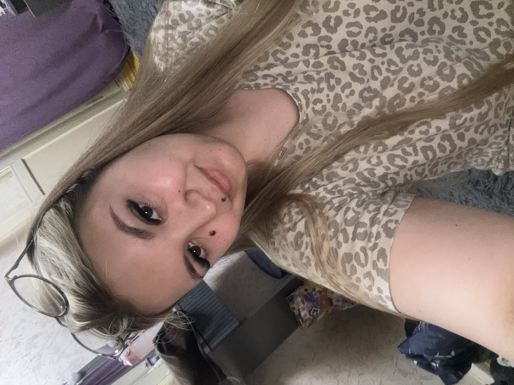 A young woman with blonde hair and glasses perched on her head is taking a selfie indoors. She is wearing a light-colored shirt with a leopard print. Her face is angled towards the camera, and she has eye makeup and two moles visible on her face. The background appears to be a room with a purple curtain and other furniture. There is no indication of the location being in Большой Букор, Russia.