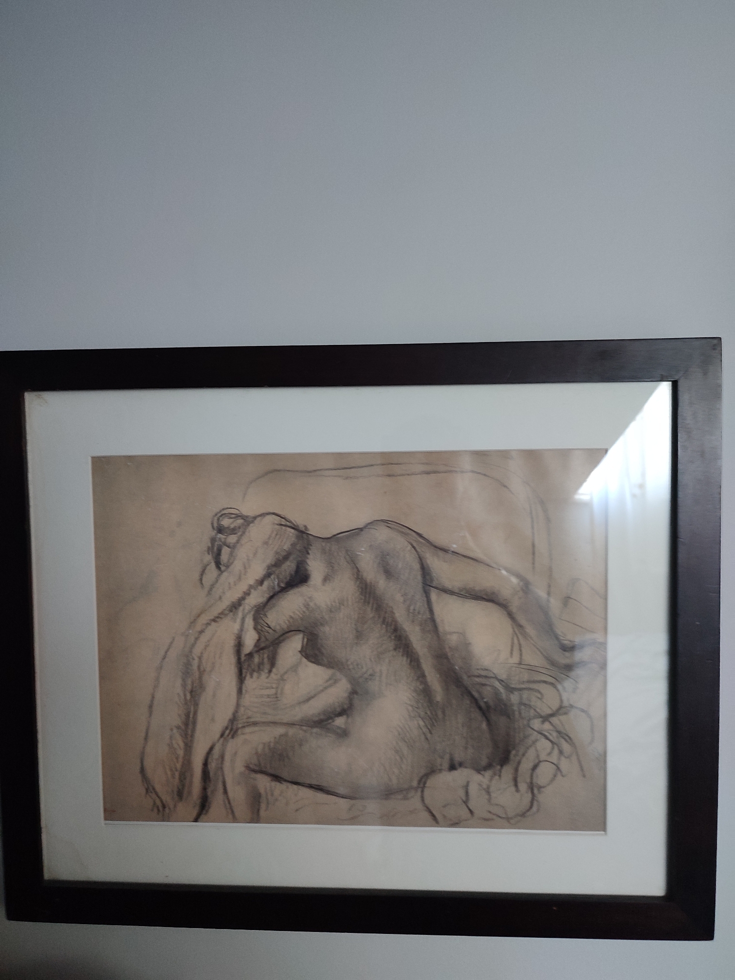 A framed charcoal drawing of a nude female figure is hanging on a light gray wall. The drawing depicts a woman from behind, seated and leaning forward, with her head bowed and hair draped over her arms. The charcoal lines are loose and expressive, with areas of shading to define the form. The drawing is matted in white and encased in a dark brown frame. There is a noticeable glare on the glass of the frame, obscuring some details. The overall impression is a study of the human form with a focus on movement and texture. The image does not contain any information about its location in Üsküdar, Turkey.