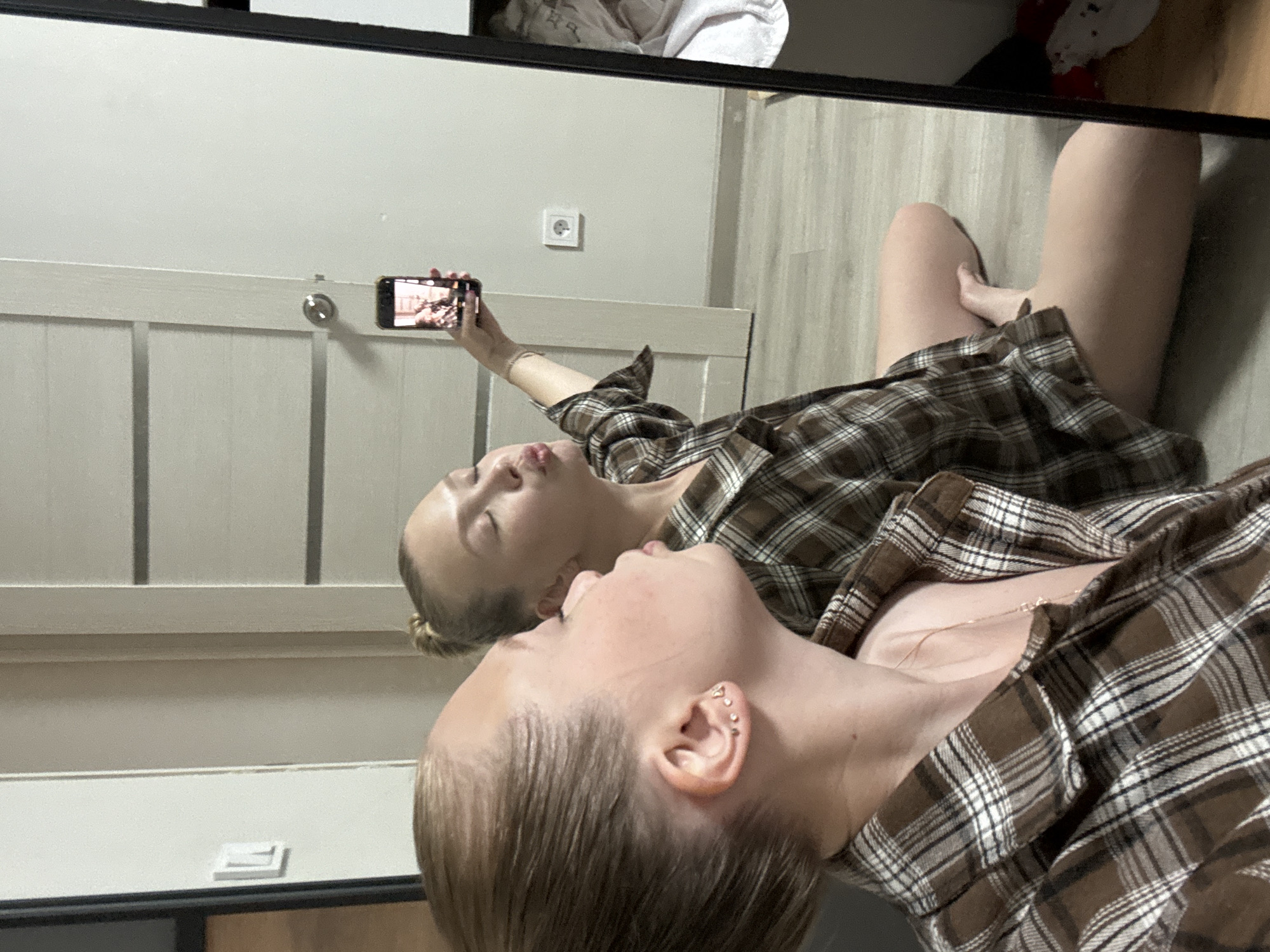 A young woman with blonde hair pulled into a bun is taking a selfie in a mirror. She is wearing a brown and white plaid button-down shirt. Her lips are pursed as she holds up her phone to take the picture. She is sitting on the floor of a room. In the background, a door and a light switch are visible on the wall. The floor appears to be made of wood-look laminate. The location is not specified in Chaikovsky, Russia.
