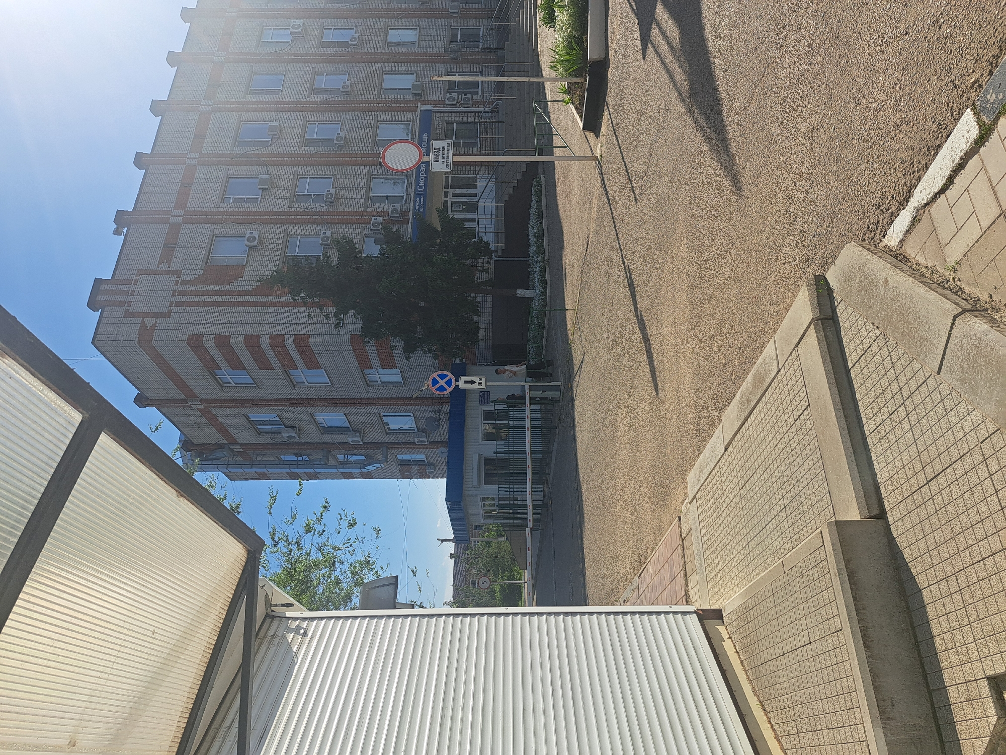 The image presents an outdoor scene in Leningradskaya, Russia. The perspective is from street level, looking towards a multi-story building constructed of gray and red brick. The building has numerous windows with air conditioning units beneath many of them. Street signs are visible in front of the building, including a red circle indicating a restricted area, and a blue sign with a white cross. A tree partially obscures the building's facade. A paved road occupies the upper right portion of the image. In the foreground, there's a tiled concrete wall and a corrugated metal roof of a structure. The sky is blue with sunlight. No people are visible in the image.