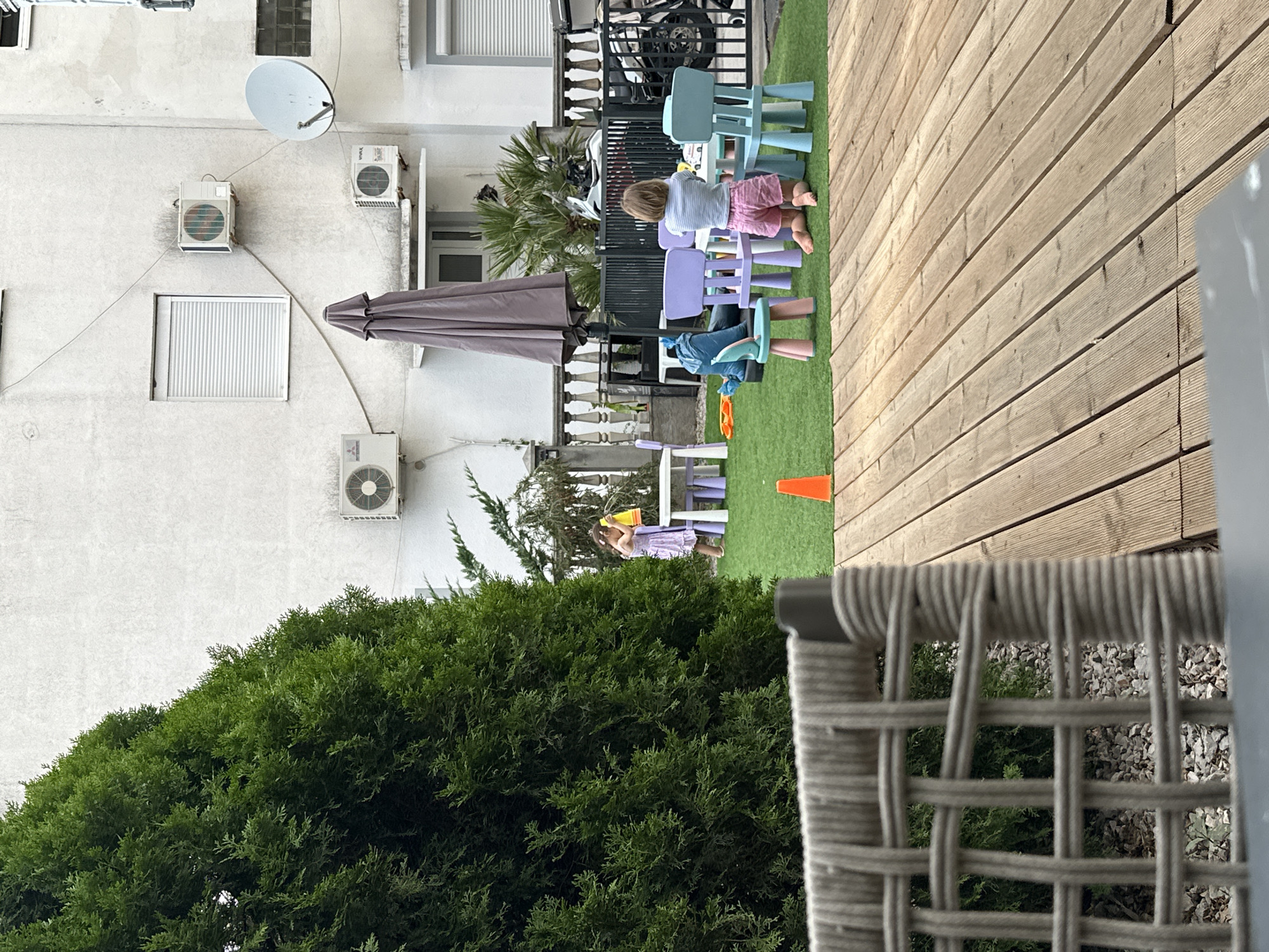 The image shows an outdoor scene in Budva, Montenegro, likely during daytime. In the foreground, a portion of a woven outdoor chair is visible. Behind the chair, a lush green hedge takes up the lower left of the frame. To the right of the hedge, a wooden deck with a diagonal plank pattern dominates the upper right portion of the image.
In the middle ground, a small group of children are playing on an artificial turf area. One child, wearing pink shorts and a striped shirt, is lying on their stomach on the grass, facing away from the camera, towards a collection of colorful plastic chairs. Another child, in a light purple dress, is standing and appears to be holding a yellow object, possibly a bucket or toy. Several other small chairs, in shades of purple and blue, are scattered around the children. An orange traffic cone is also visible on the grass.
In the background, a white building with several air conditioning units and a satellite dish mounted on its exterior is visible. An open window with white shutters is also present. A closed umbrella is leaning against the building. A motorcycle is parked behind a black metal fence. The overall atmosphere suggests a casual, recreational setting for children.