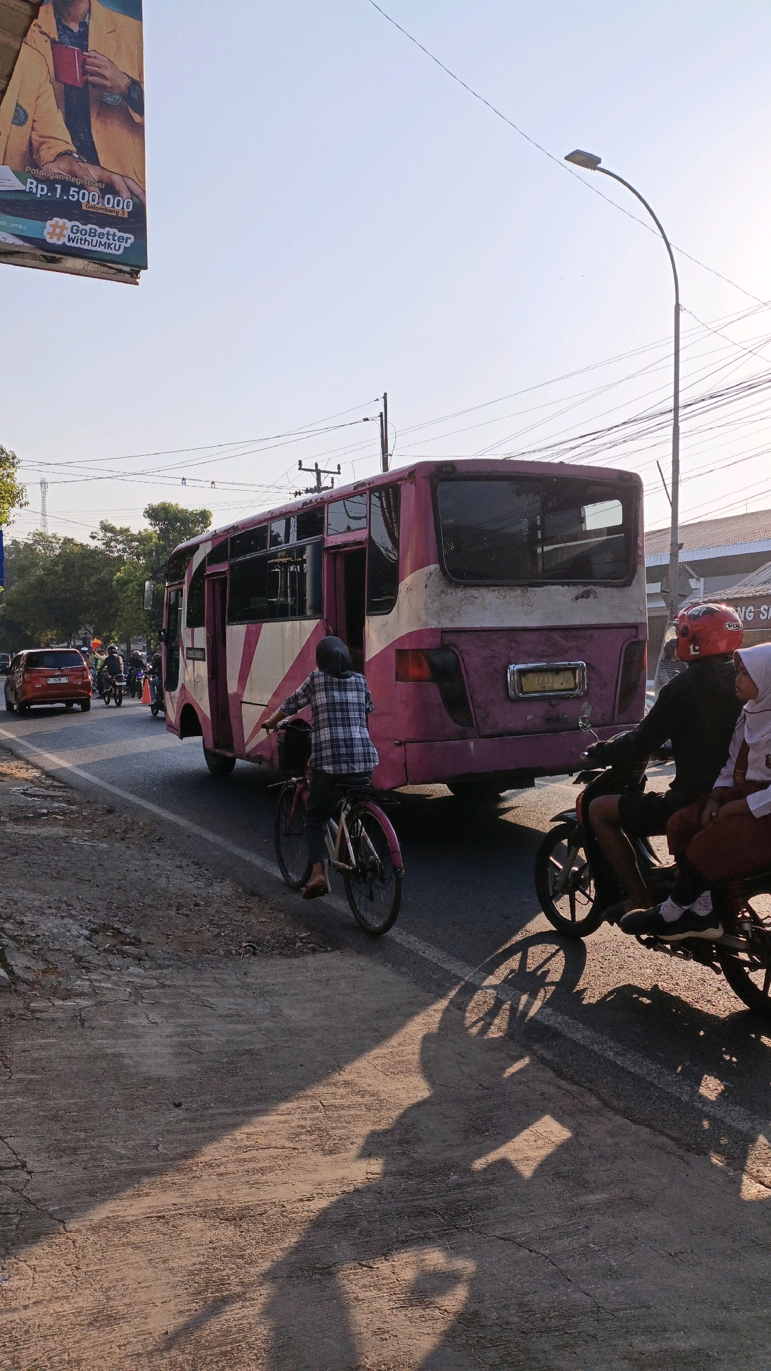 A pink and white bus is driving on a paved road in Kuwasen, Indonesia. The bus is in motion, and its doors are open. A person is riding a bicycle on the left side of the bus. Two people are on a motorcycle on the right side of the bus. The motorcycle is also in motion. There are trees and buildings in the background. The sun is shining, and there are shadows on the road.