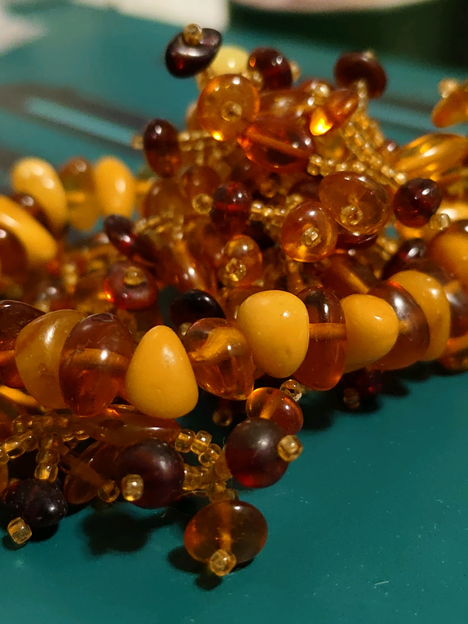 The image shows a close-up, macro view of a strand of amber beads. The beads are various shades of yellow, orange, and dark red, with some appearing translucent and others opaque. They are strung together with small, golden seed beads. The beads are clustered and overlapping, creating a textured and somewhat chaotic appearance. The background is a solid, teal-colored surface, possibly a table or countertop. There is no indication of a specific location in Rostov-on-Don, Russia, or any people or actions. The lighting appears to be artificial, with some highlights reflecting off the polished surfaces of the beads.