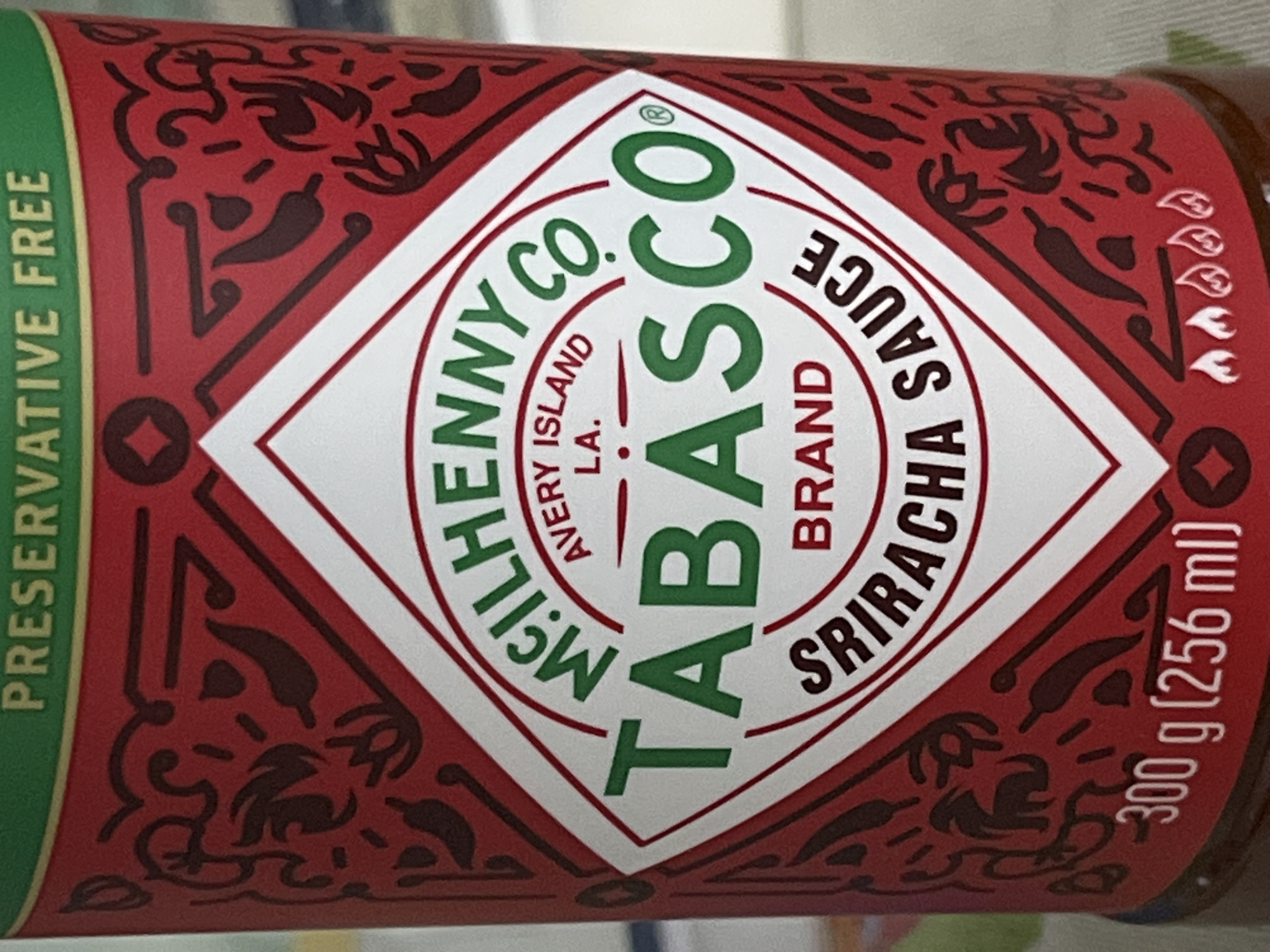 A close-up, rotated shot captures the label of a Tabasco Brand Sriracha Sauce bottle. The label is predominantly red with intricate dark red patterns featuring chili peppers and abstract swirls. A central white diamond-shaped area prominently displays the product information.
Visible text on the label includes "PRESERVATIVE FREE" in green along the left edge. The main brand identity, "MCILHENNY CO. AVERY ISLAND LA. TABASCO BRAND SRIRACHA SAUCE," is displayed in a circular green font around the "TABASCO" logo, which is vertically oriented. Below this, "SRIRACHA SAUCE" is horizontally aligned. Further to the right, the weight and volume are indicated as "300 g (256 ml)." The background is blurred, suggesting a shallow depth of field, bringing the product label into sharp focus. The overall impression is a vibrant and detailed product advertisement.