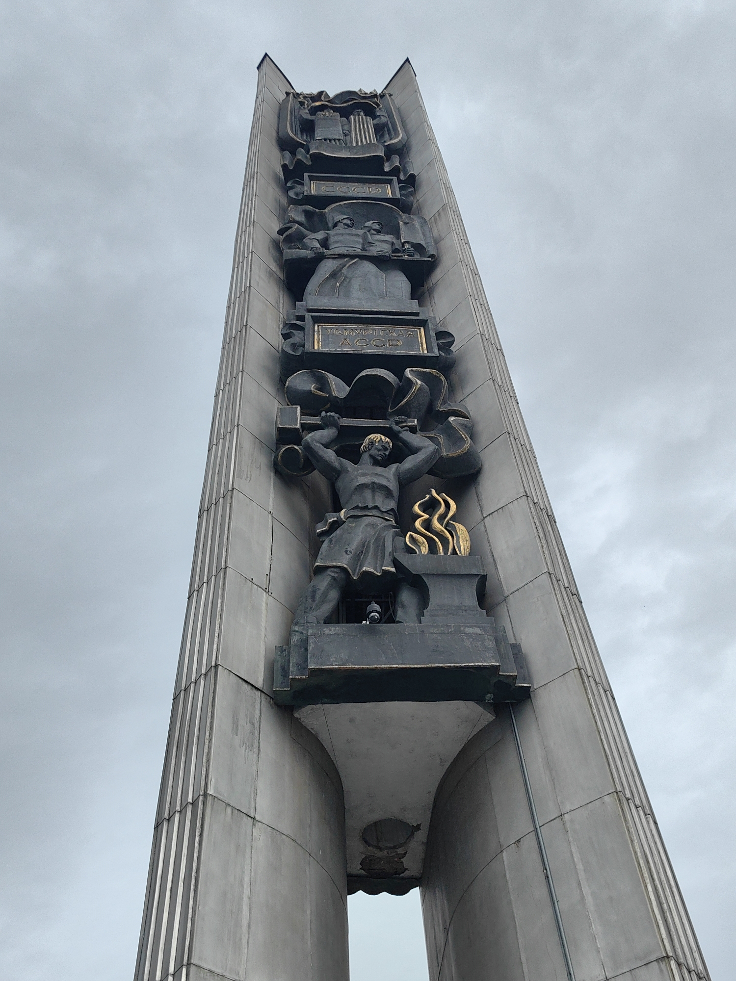 A low-angle shot captures a tall, grey monument in Izhevsk, Russia, set against an overcast sky. The monument features four distinct sculptural tiers. The top tier shows two figures draped in classical garb. The next tier shows two figures carrying a banner. The third tier shows a muscular figure holding an industrial part, with golden flames to the figure's side. The base of the monument features two supports on either side of an archway.