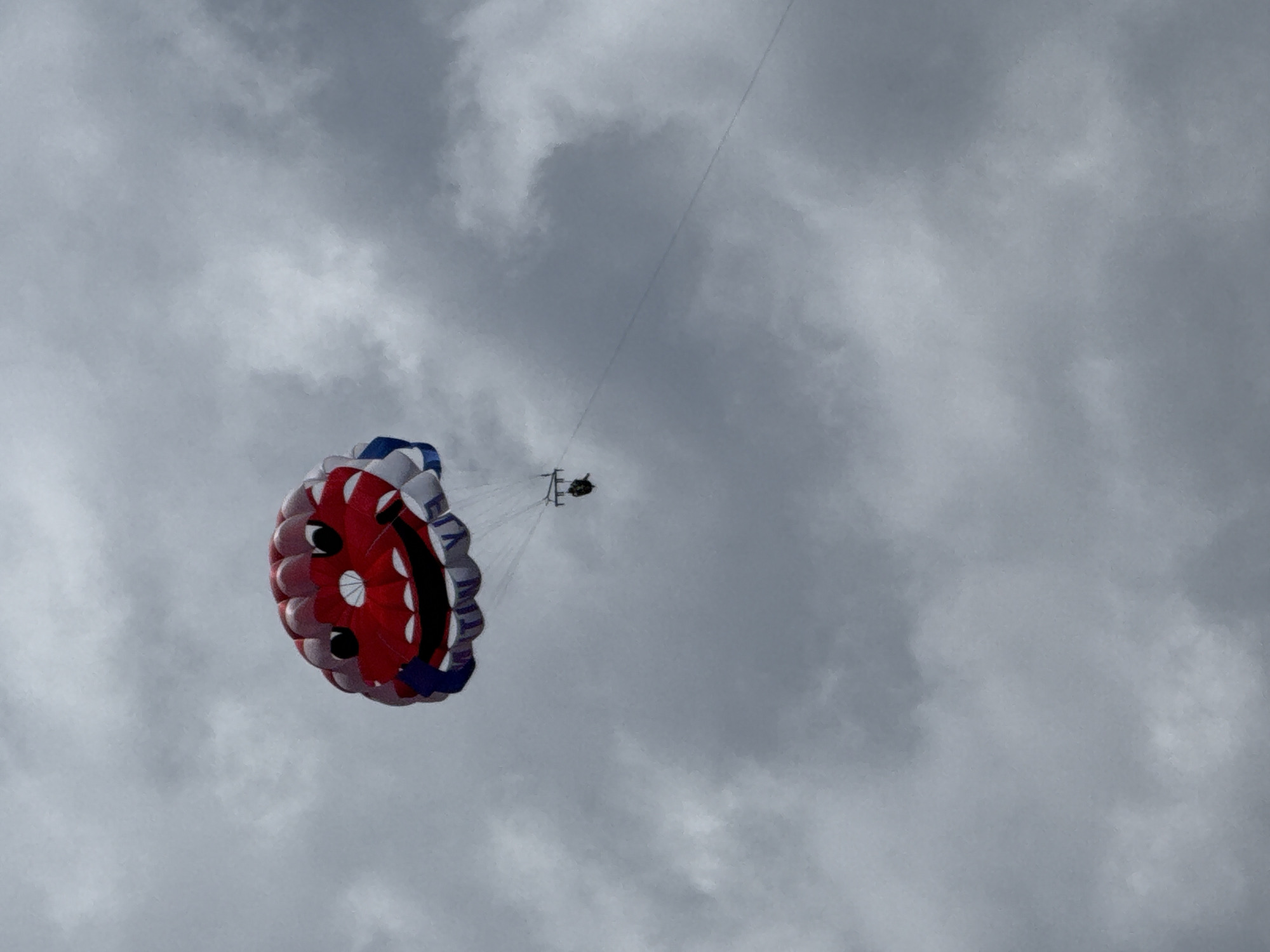Against a backdrop of a dense, overcast sky, a parasailer soars high above. The scene, likely set in Budva, Montenegro, captures the parasail in mid-air, connected by a thin line disappearing towards the upper right, implying a tow from a boat out of frame.
The parasail itself is vibrantly colored, primarily red and white with blue accents. A notable detail is the design on its underside, which strikingly resembles a smiling face, complete with dark, rounded shapes for eyes and a curved line for a mouth. Suspended beneath the large canopy, the dark, small silhouette of a person or people in a harness is visible, indicating the individual(s) enjoying the aerial activity.
The sky is filled with heavy, grey clouds, creating an atmosphere of soft, diffused light typical of an overcast day. Despite the lack of direct sunlight, it is clearly daytime. Faint text can be discerned on the white and blue sections of the parasail, appearing to begin with "FLY" followed by other unreadable letters, likely a brand or company name. The image is taken from a ground-level perspective looking upwards, emphasizing the height and expanse of the sky.