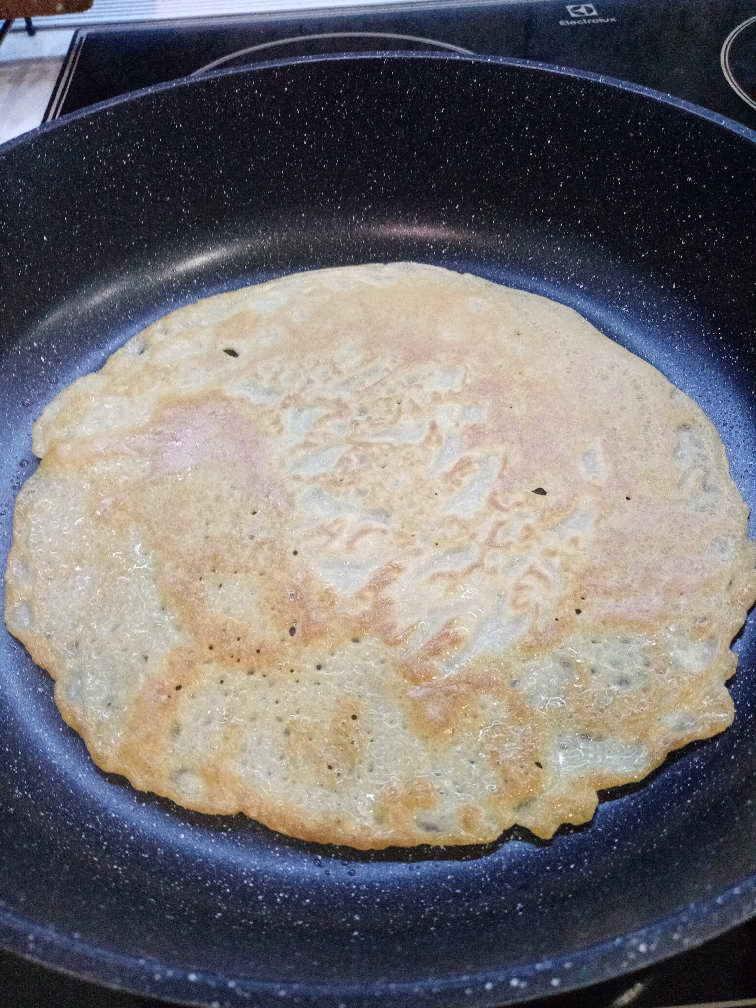 The image shows a round, light brown crepe cooking in a non-stick pan with a speckled, dark blue/black surface. The pan is on an electric stovetop with the brand name "Electrolux" visible in the background. There are no people visible. The location cannot be determined to be in Izhevsk, Russia based on the image.