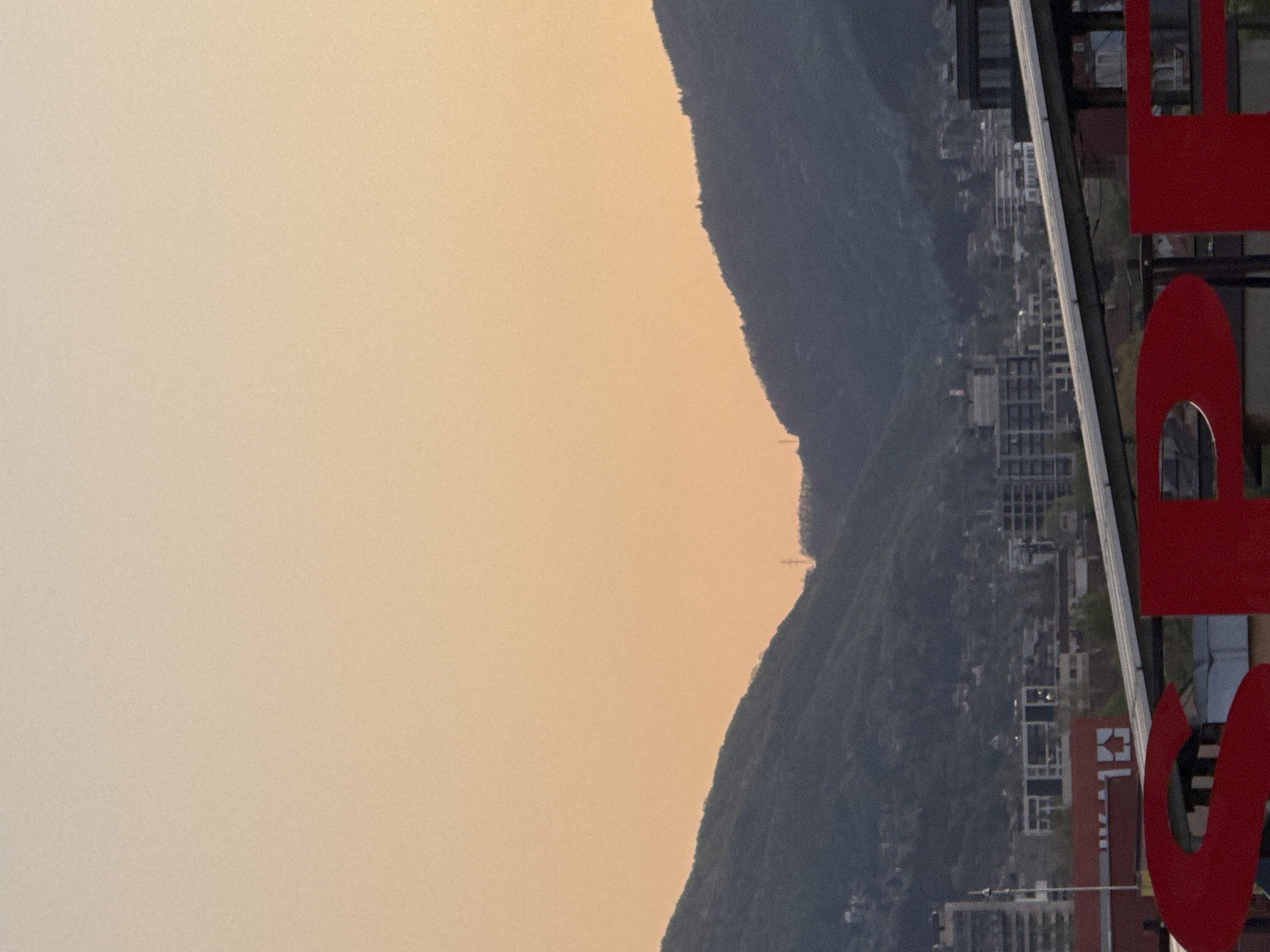 The image presents an outdoor scene with a large mountain or hill range in the background. The sky has a light orange/yellow hue, suggesting either sunrise or sunset. A city or town is nestled on the slopes of the mountain, with various buildings visible. On the right side of the image, a part of a red sign is visible, obstructing the view. Based on the architectural style and the prominence of a Canadian flag-like logo on a building, the image is likely taken from somewhere in Sofia, Bulgaria.