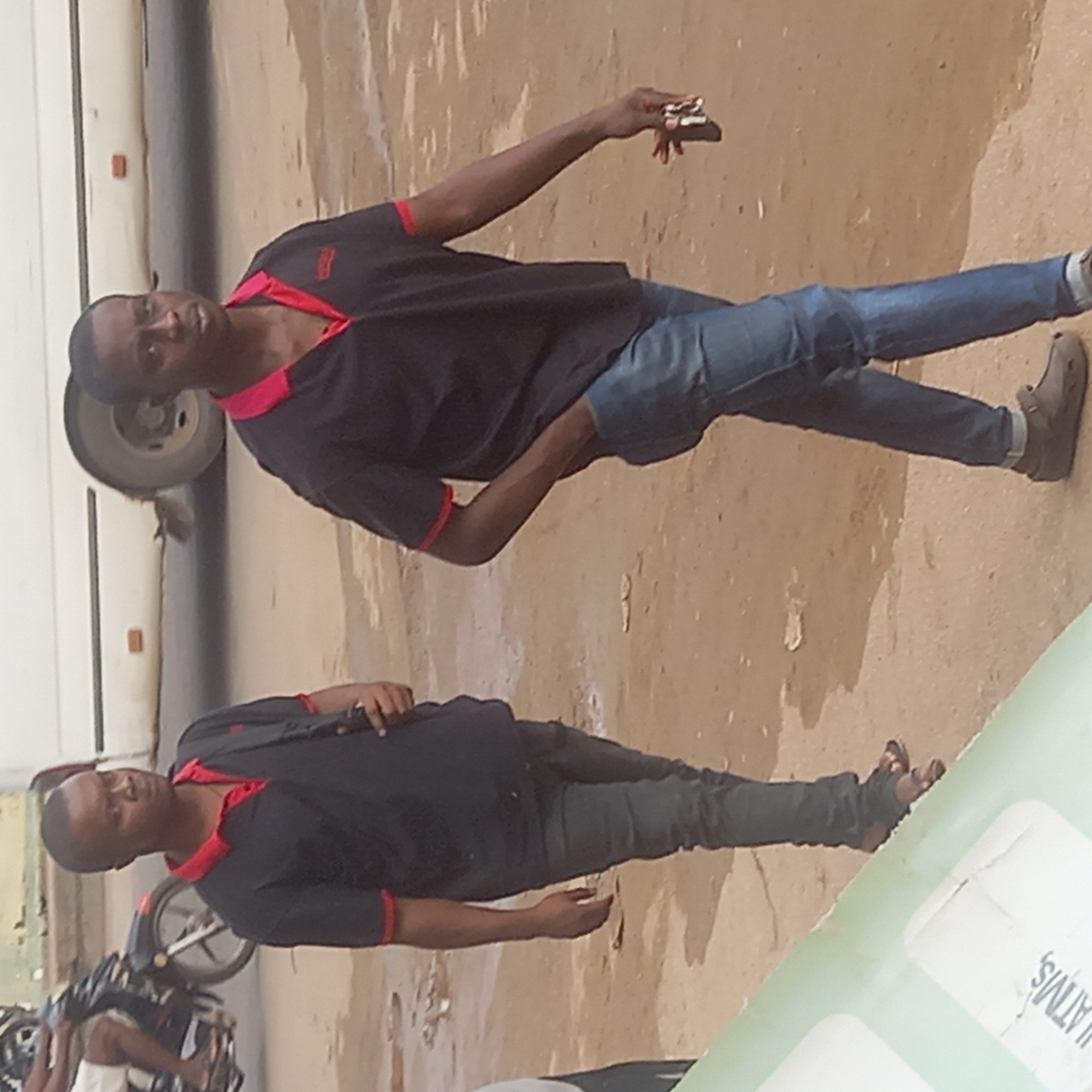 The image depicts a man in the city of Sardauna, Nigeria. He is wearing a black polo shirt with red trim, blue jeans, and brown sandals. He is standing near a white vehicle and a parked motorcycle. He is holding a small object in his raised hand. The background shows a sandy or dirt surface, typical of a street in Sardauna. A partial view of what seems to be a sign is on the right side.